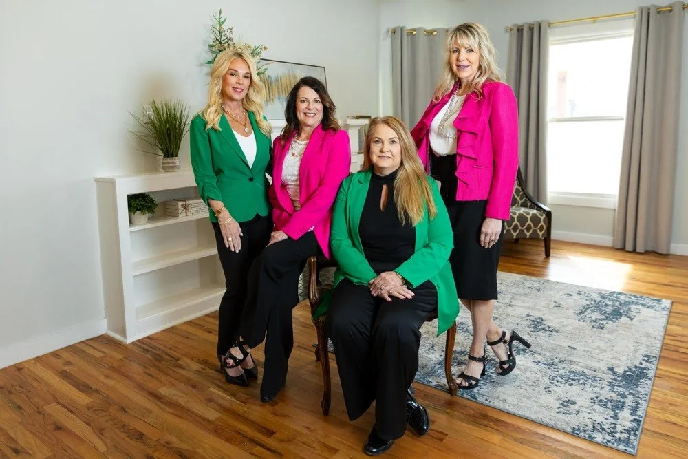 Four women dressed in colorful blazers, standing and sitting in a bright, modern living room with hardwood floors and a rug, smiling for a group photo.