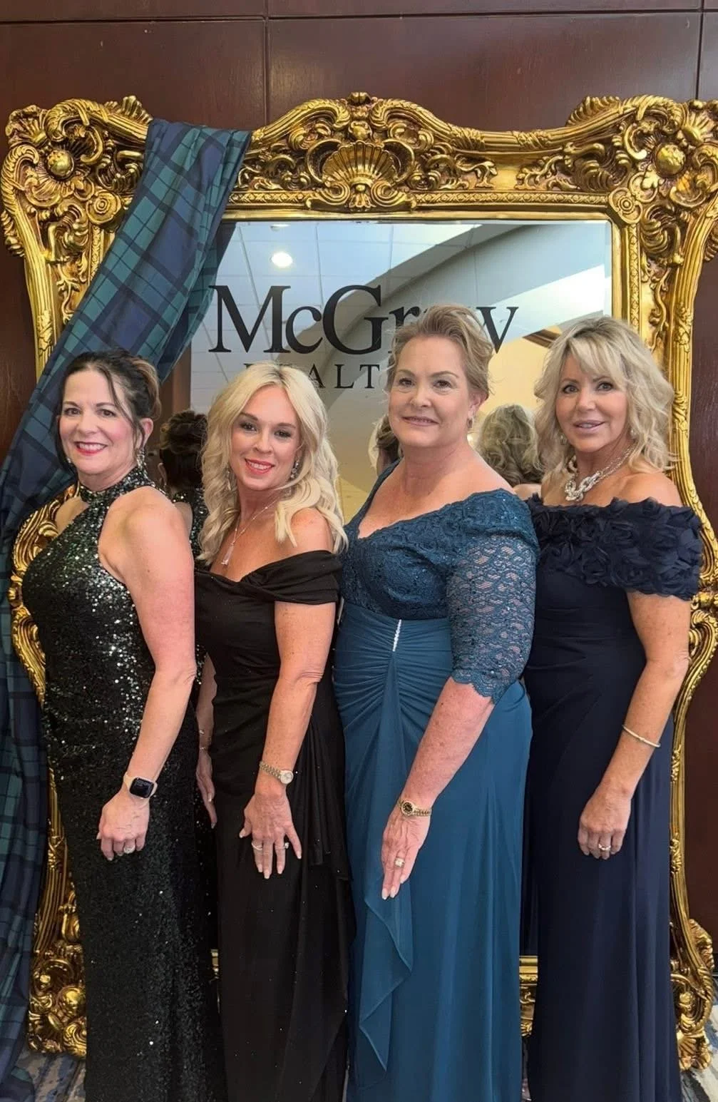 Four women in formal evening dresses standing in front of a gold ornate mirror with the word 'McGraw' visible in the reflection, at an event or celebration.