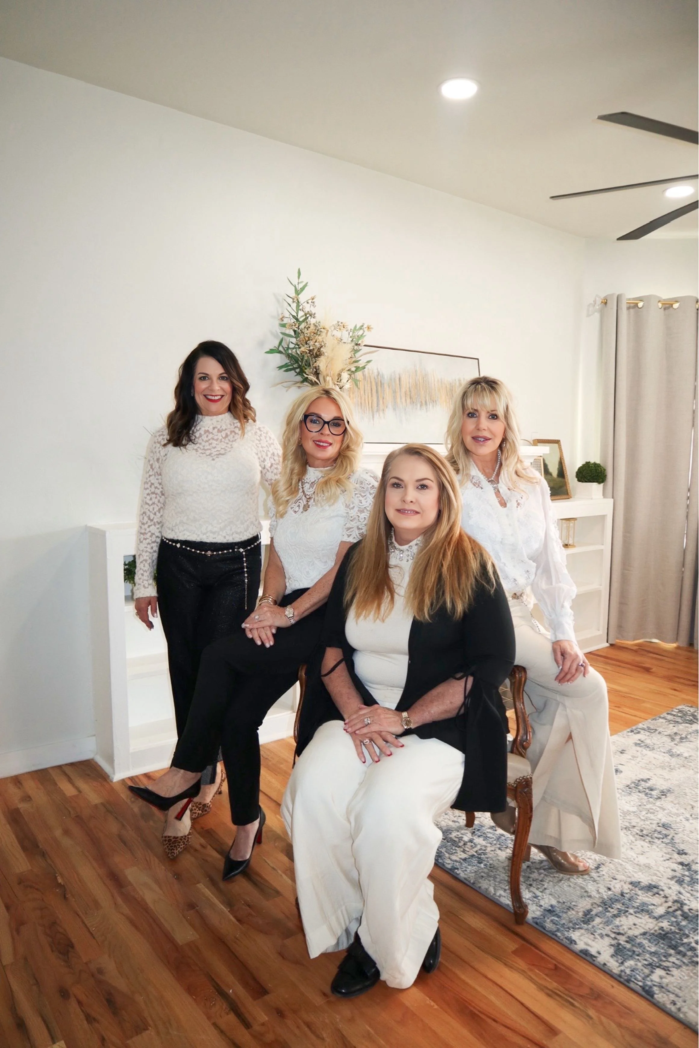 A group of five women in elegant attire posing together in a well-lit, modern living room with hardwood floors, white walls, and minimal decor, including a painting, a plant, and curtains.