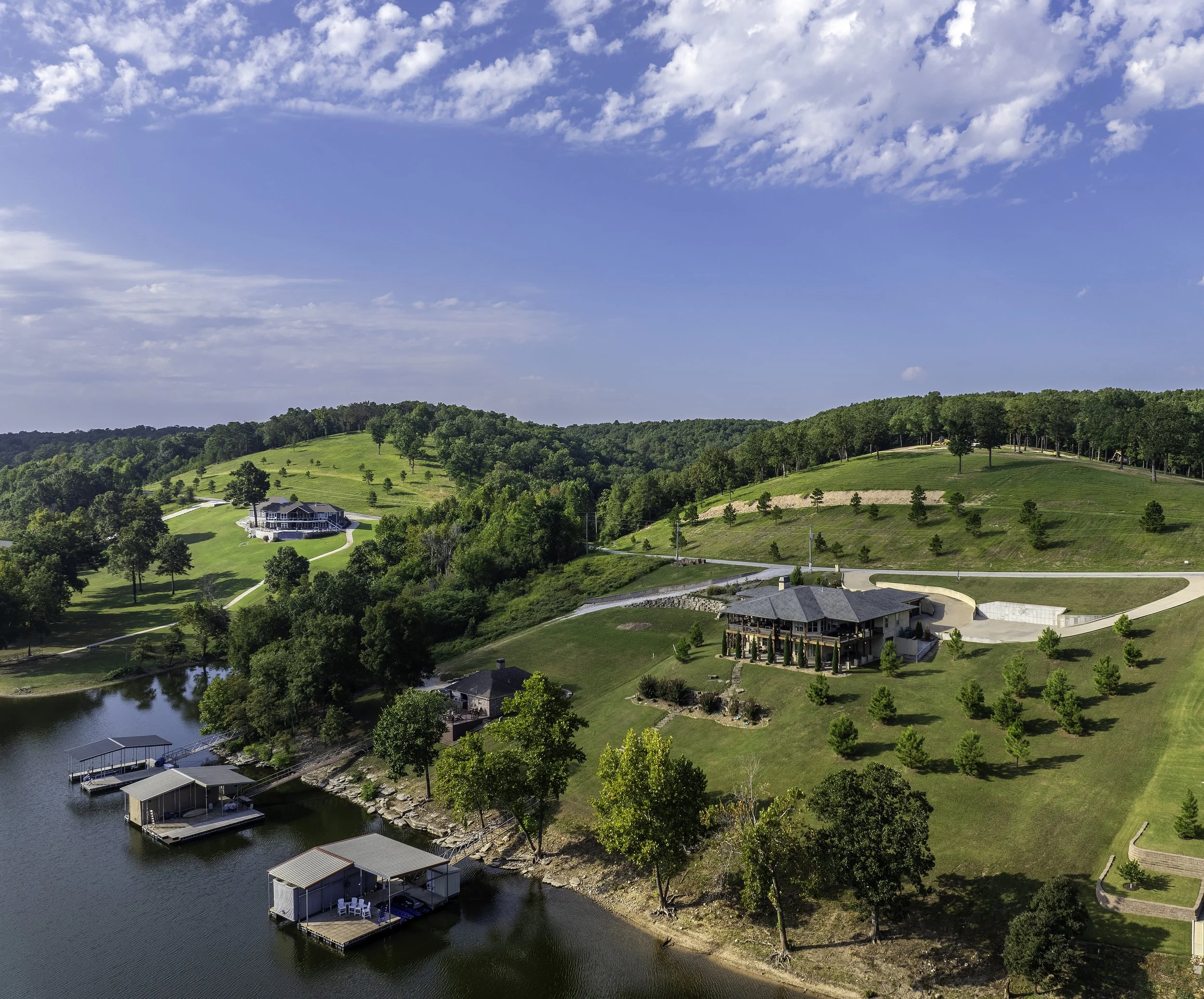 A scenic view of a lake with docked boat houses, green rolling hills, scattered trees, and a clear blue sky with white clouds.