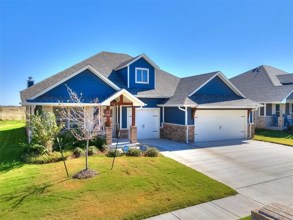 A modern, blue-colored house with a triple-car garage, front porch, small trees, and well-maintained lawn under a bright blue sky.