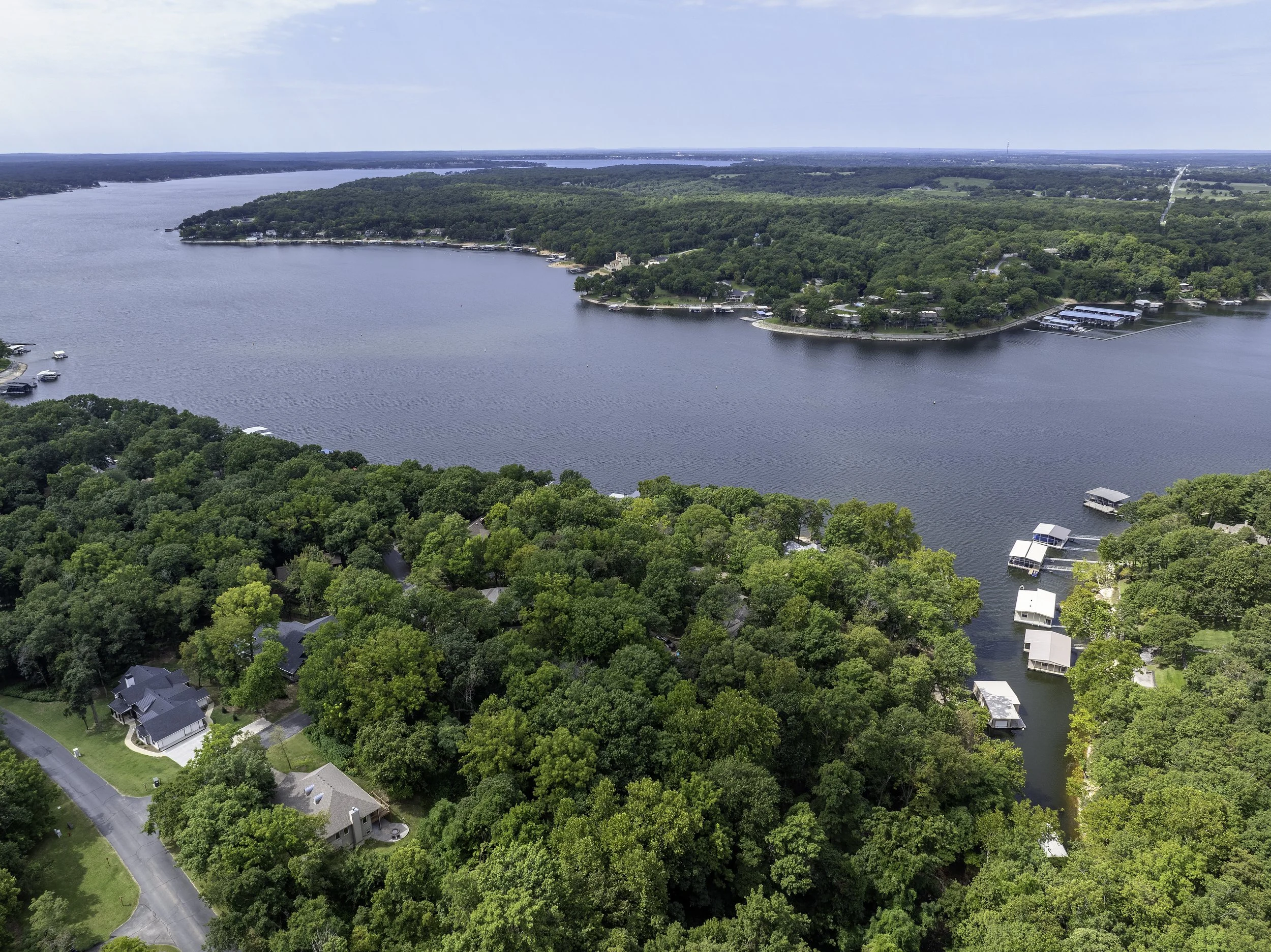 Aerial view of a large lake surrounded by lush green trees and residential houses, with some boat docks along the shoreline, under a partly cloudy sky.
