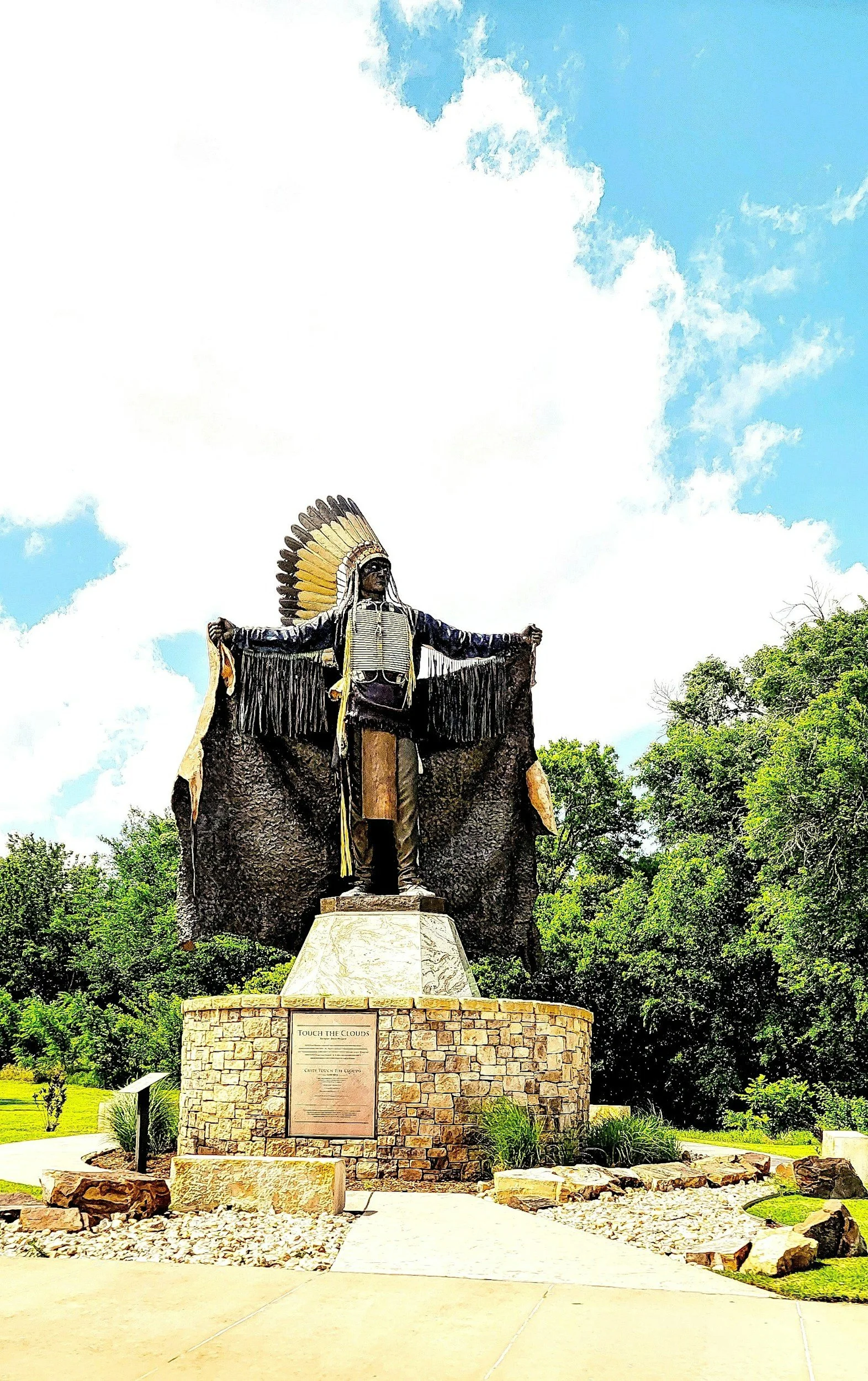 Statue of a Native American with a feathered headdress, standing with arms outstretched against a sky with clouds. The statue is on a brick pedestal with a plaque and is surrounded by greenery and rocks.