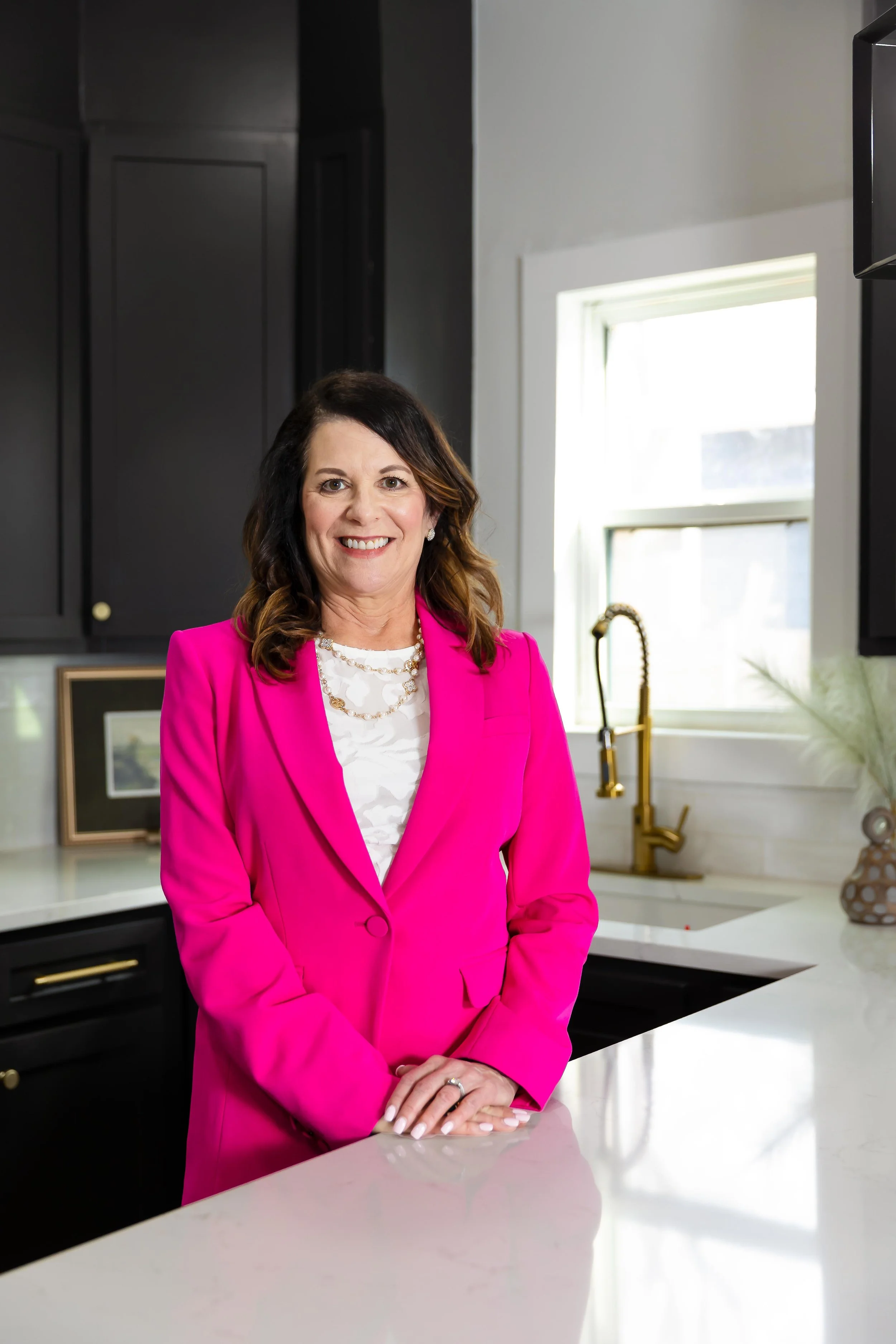 A woman with shoulder-length brown hair and a bright smile, wearing a vibrant pink blazer and white blouse, standing in a modern kitchen with black cabinets, white countertops, and a window in the background.