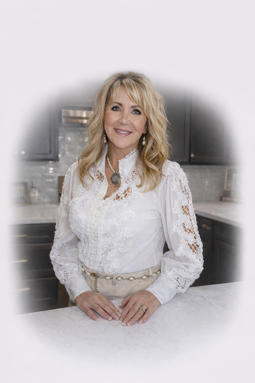 A woman with blonde wavy hair and blue eyes, wearing a white lace blouse, standing in a modern kitchen with dark cabinets and marble countertops, smiling at the camera.