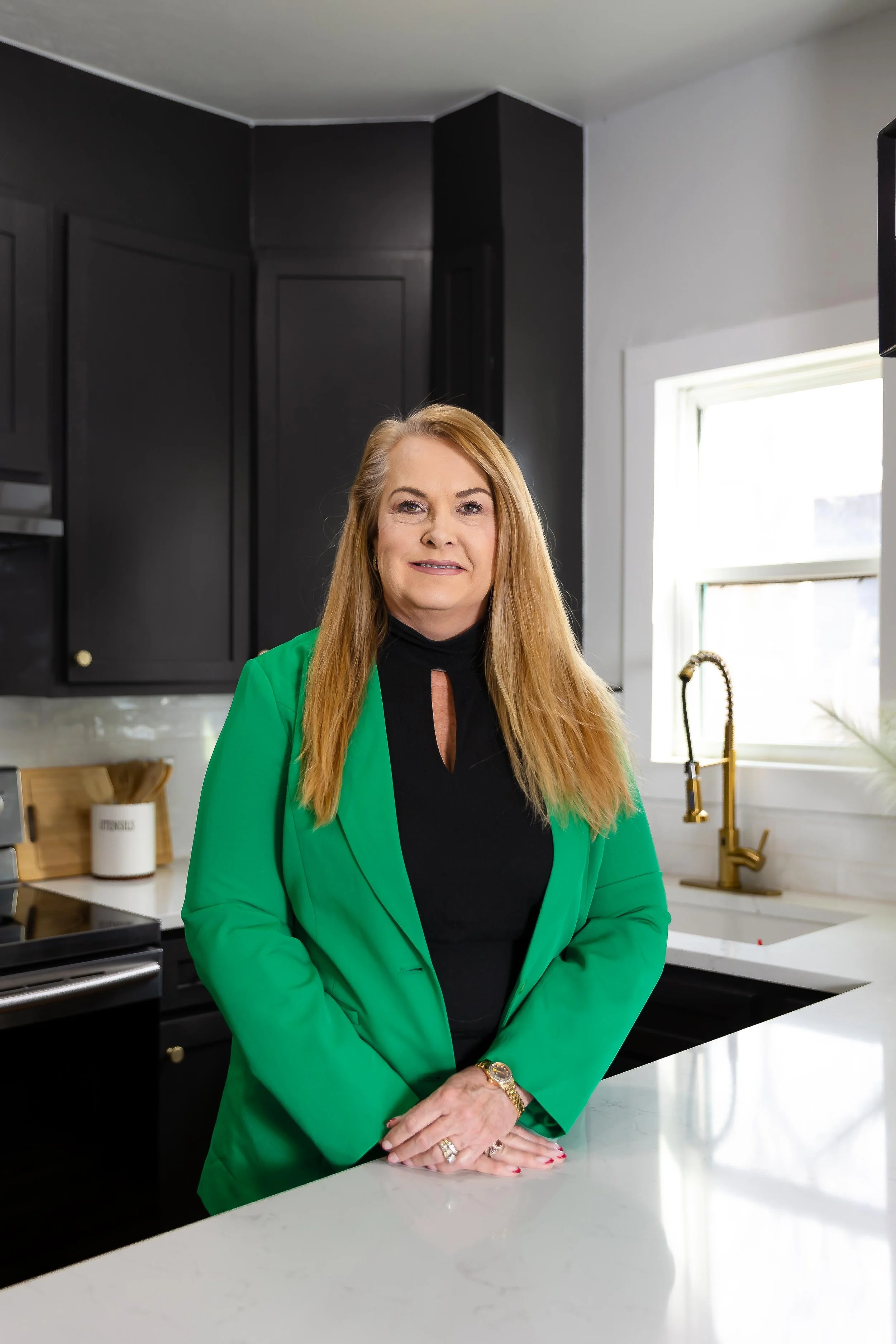 A woman with long blonde hair wearing a black top and green blazer, standing in a modern kitchen with black cabinets, a white countertop, a brass faucet, and a window with natural light.