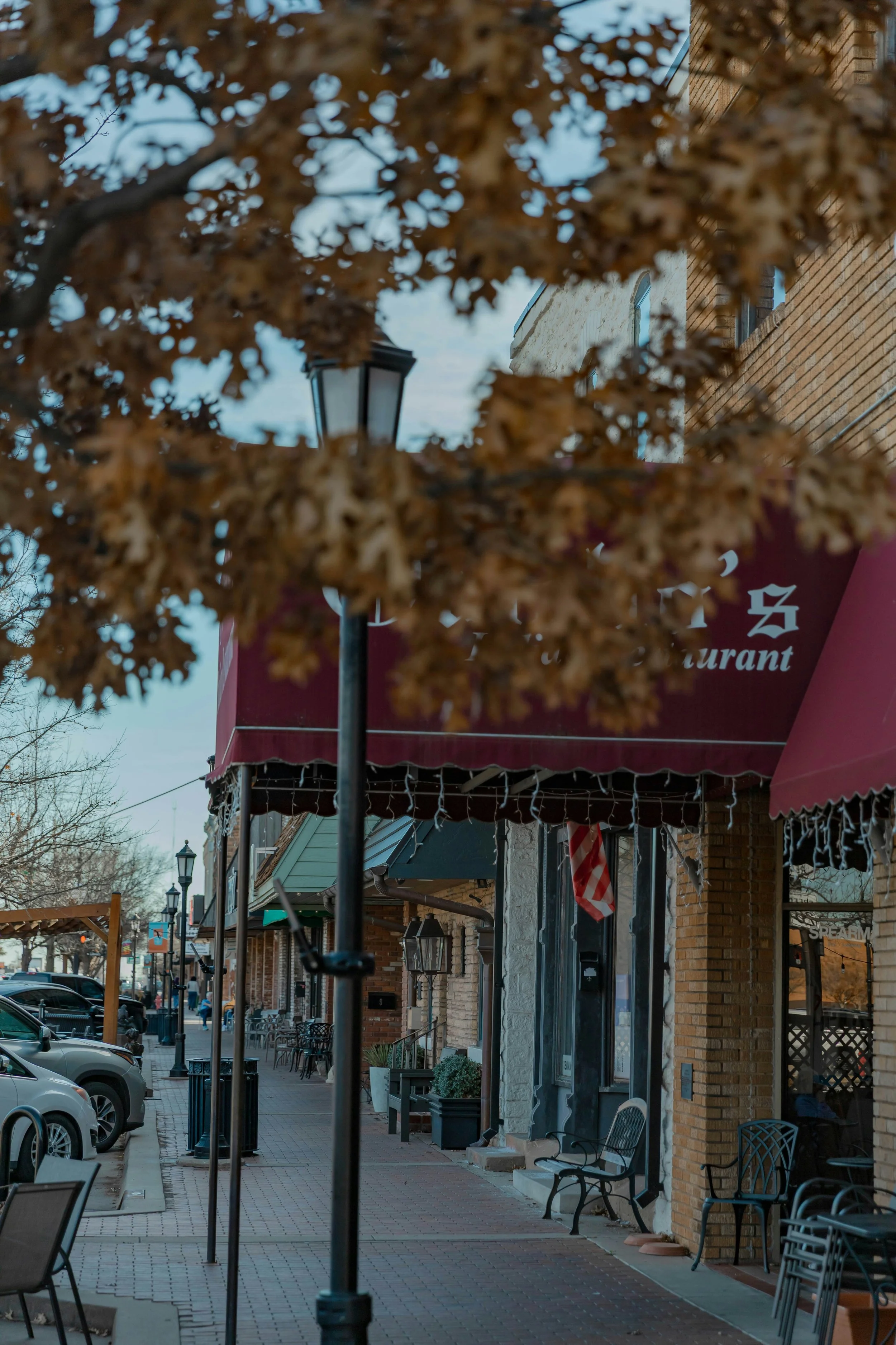 A street view taken during the day with outdoor restaurant seating, benches, and parked cars. A lamppost is visible in the foreground. Buildings with brick and stone facades line the street, and there are leafless trees along the sidewalk.
