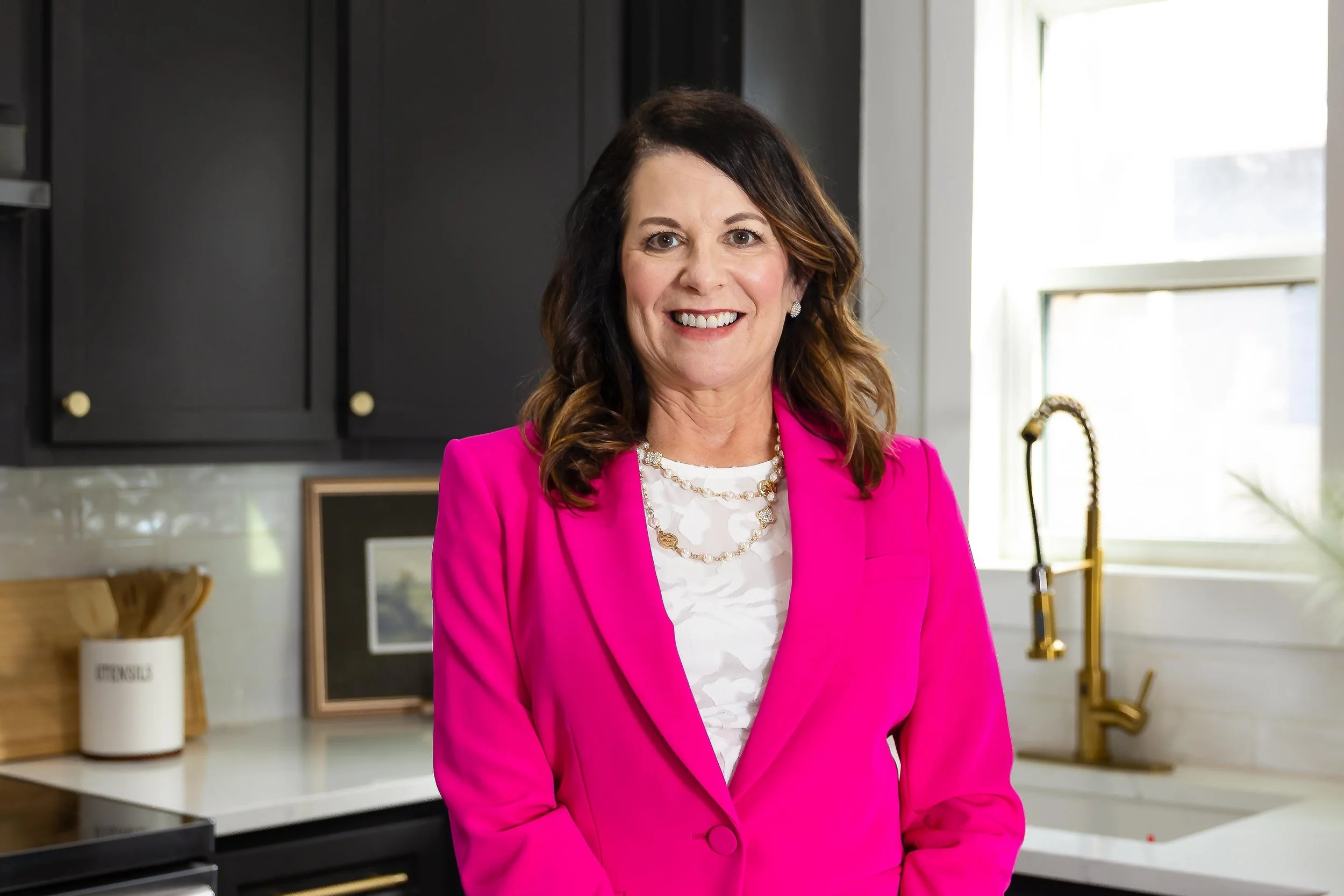 A woman with shoulder-length dark brown hair, wearing a bright pink blazer over a white blouse with a pearl necklace, smiling in a modern kitchen with black cabinets, a gold faucet, and sunlight coming through a window.