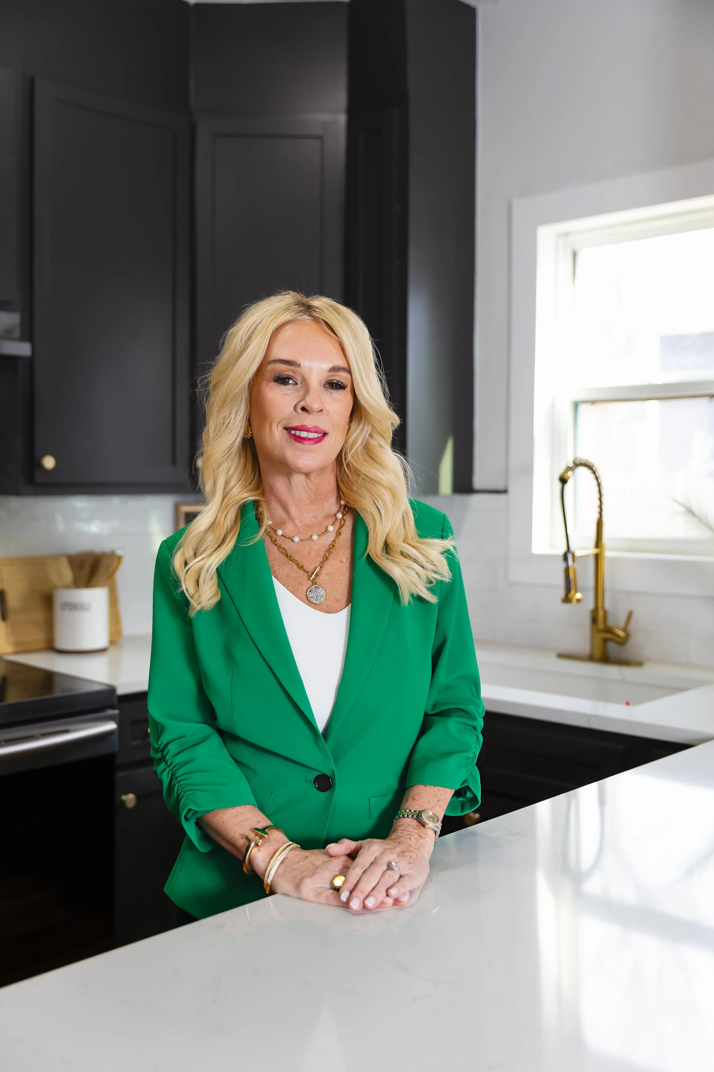 A woman with long blonde wavy hair, wearing a green blazer over a white top, standing in a modern kitchen. She has jewelry including necklaces, bracelets, and rings, and is leaning on a white countertop with her hands clasped together. The kitchen has dark cabinets, a gold faucet, and a window allowing natural light to illuminate the space.