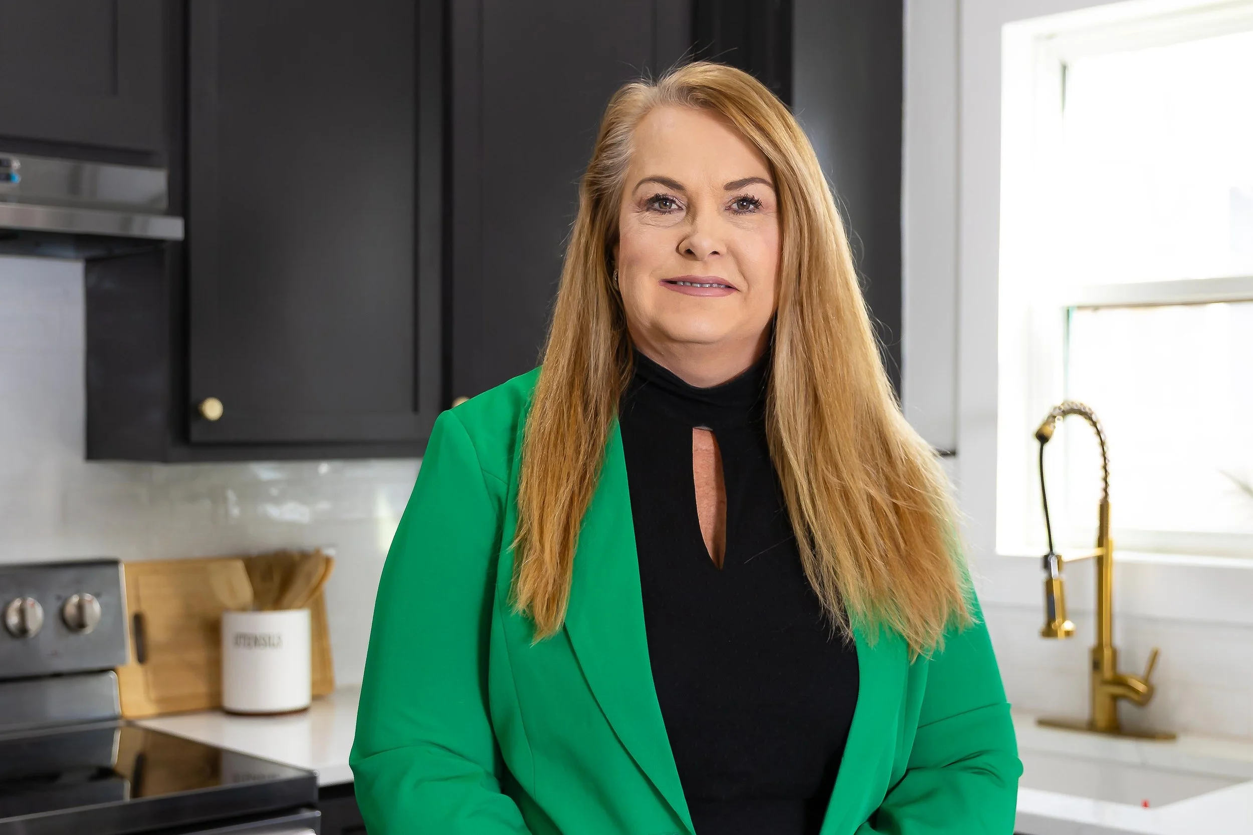 A woman with long red hair wearing a green blazer and black top, standing in a modern kitchen with black cabinets, a window, and kitchen utensils.