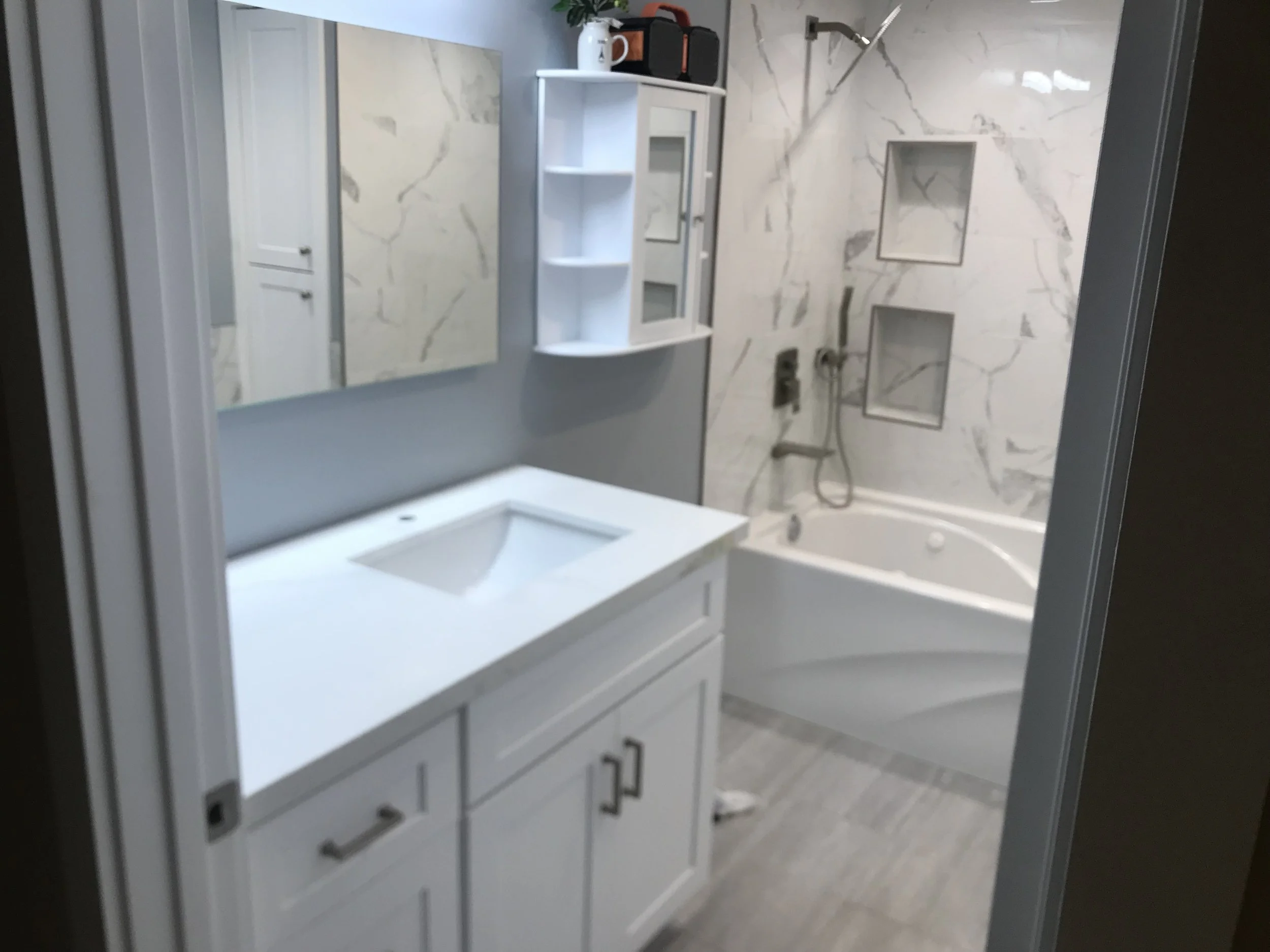A bathroom with a white vanity, a large mirror, and a shower area with marble-patterned tiles and built-in shelves.