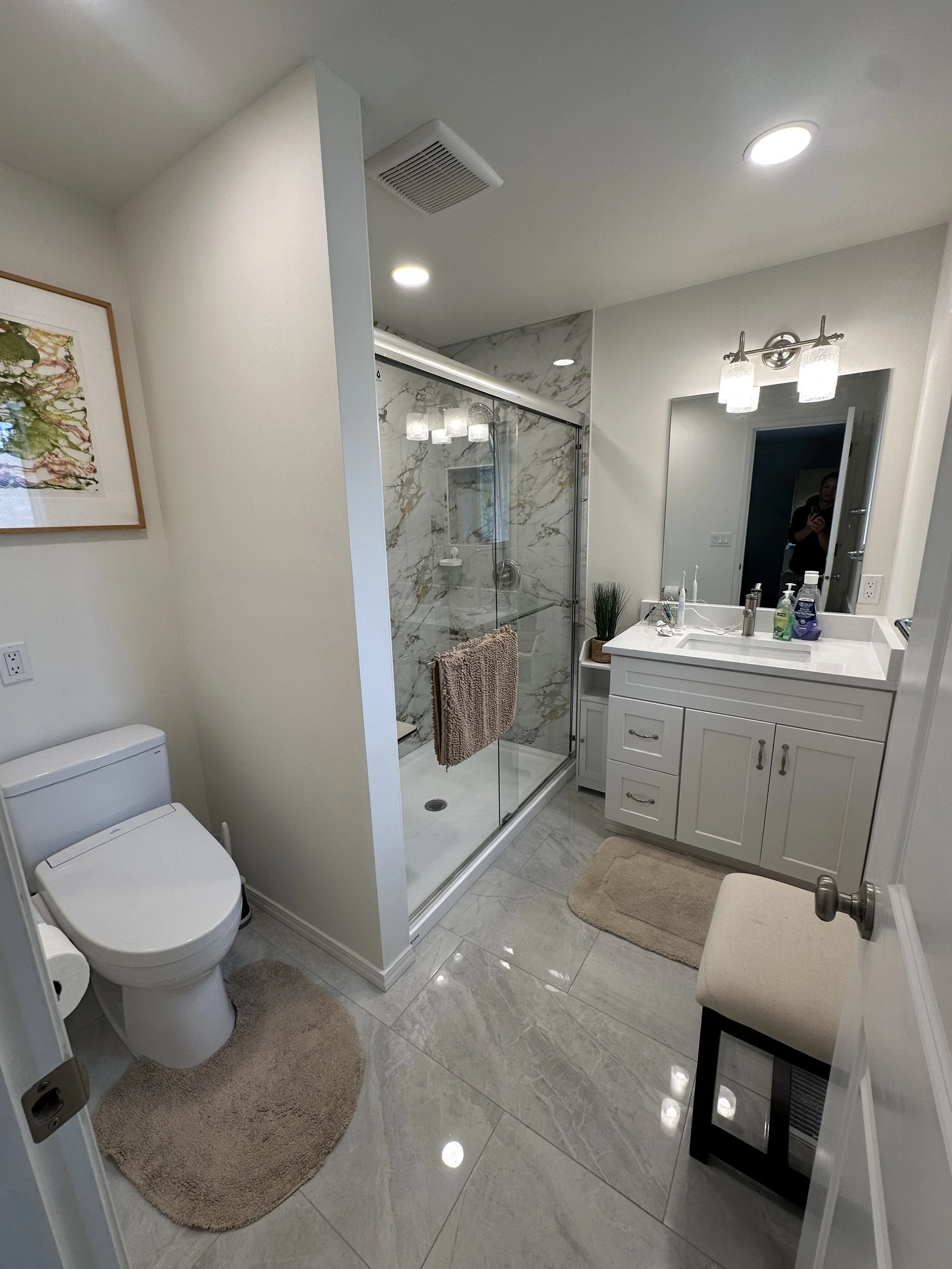 A bathroom with a toilet, a glass shower stall with marble pattern walls, a white vanity with a mirror and various toiletries, radiator, and beige rugs on the tiled floor.