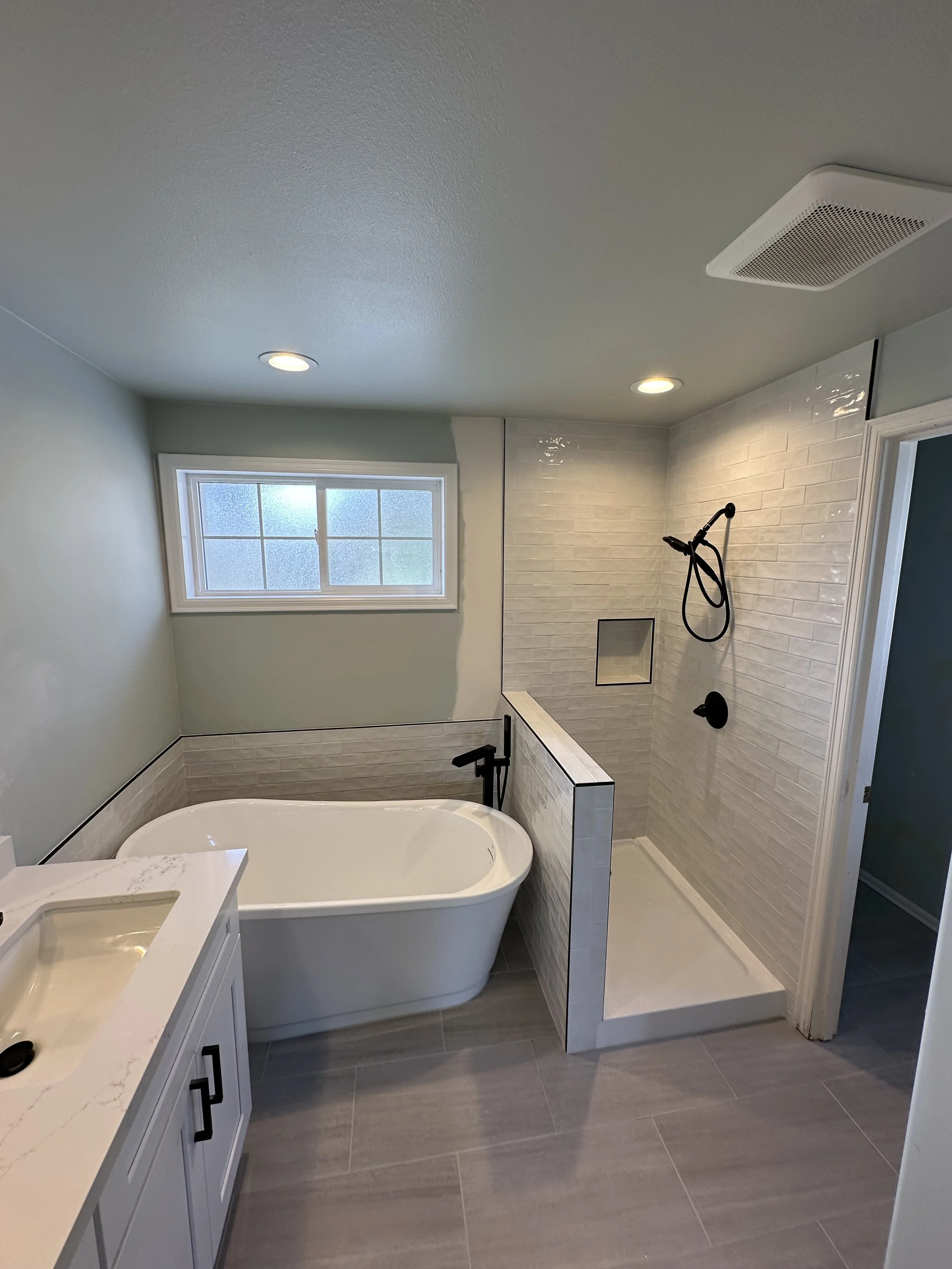 Bathroom with a bathtub, vanity with sink, window, and a walk-in shower with beige tiles and black fixtures.