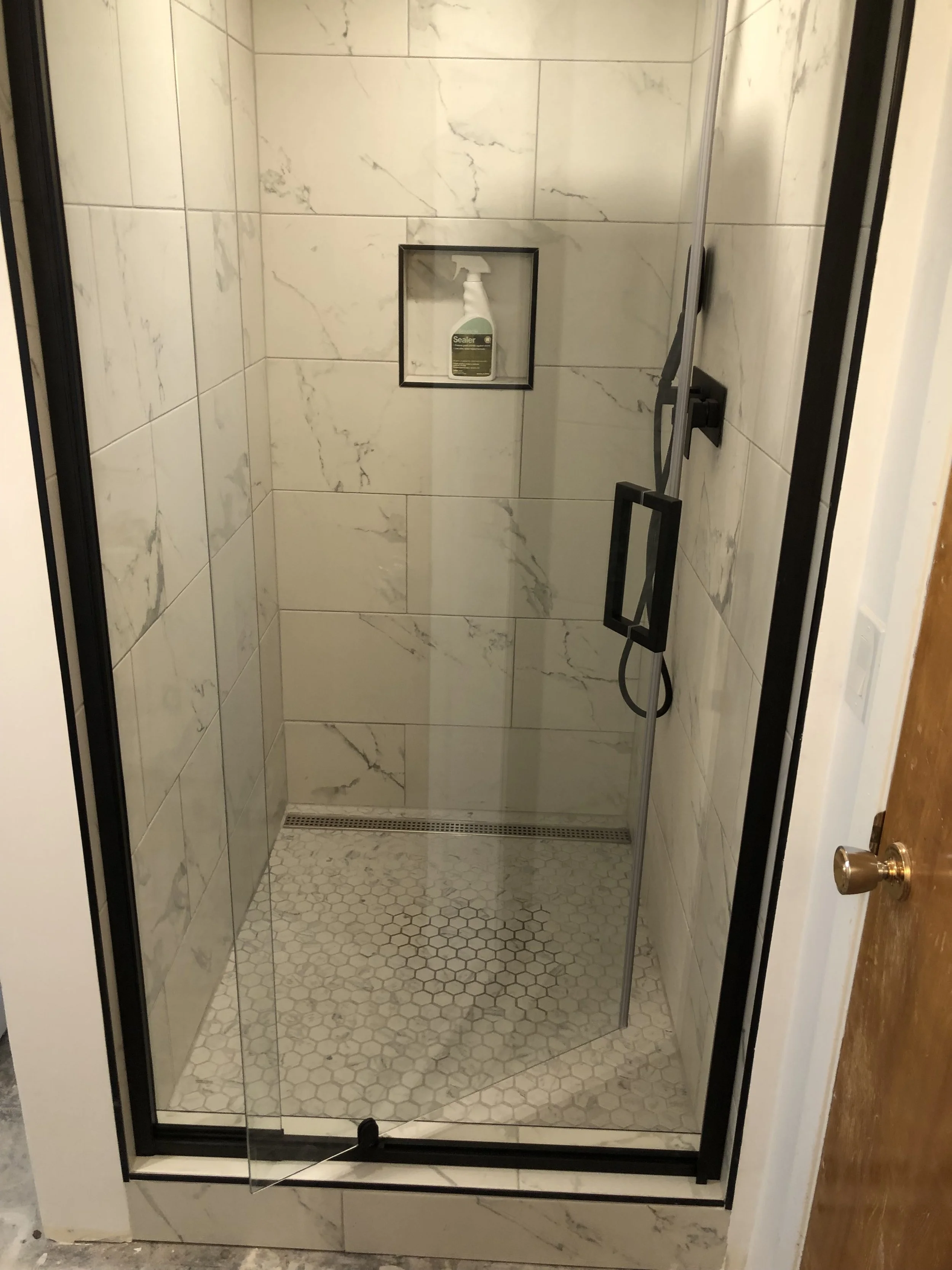 A shower with white marble tiles on the walls and small hexagonal tiles on the floor, featuring a built-in shelf with a spray bottle of cleaner, a black-framed glass door, a black shower handle, and a small drain at the base.