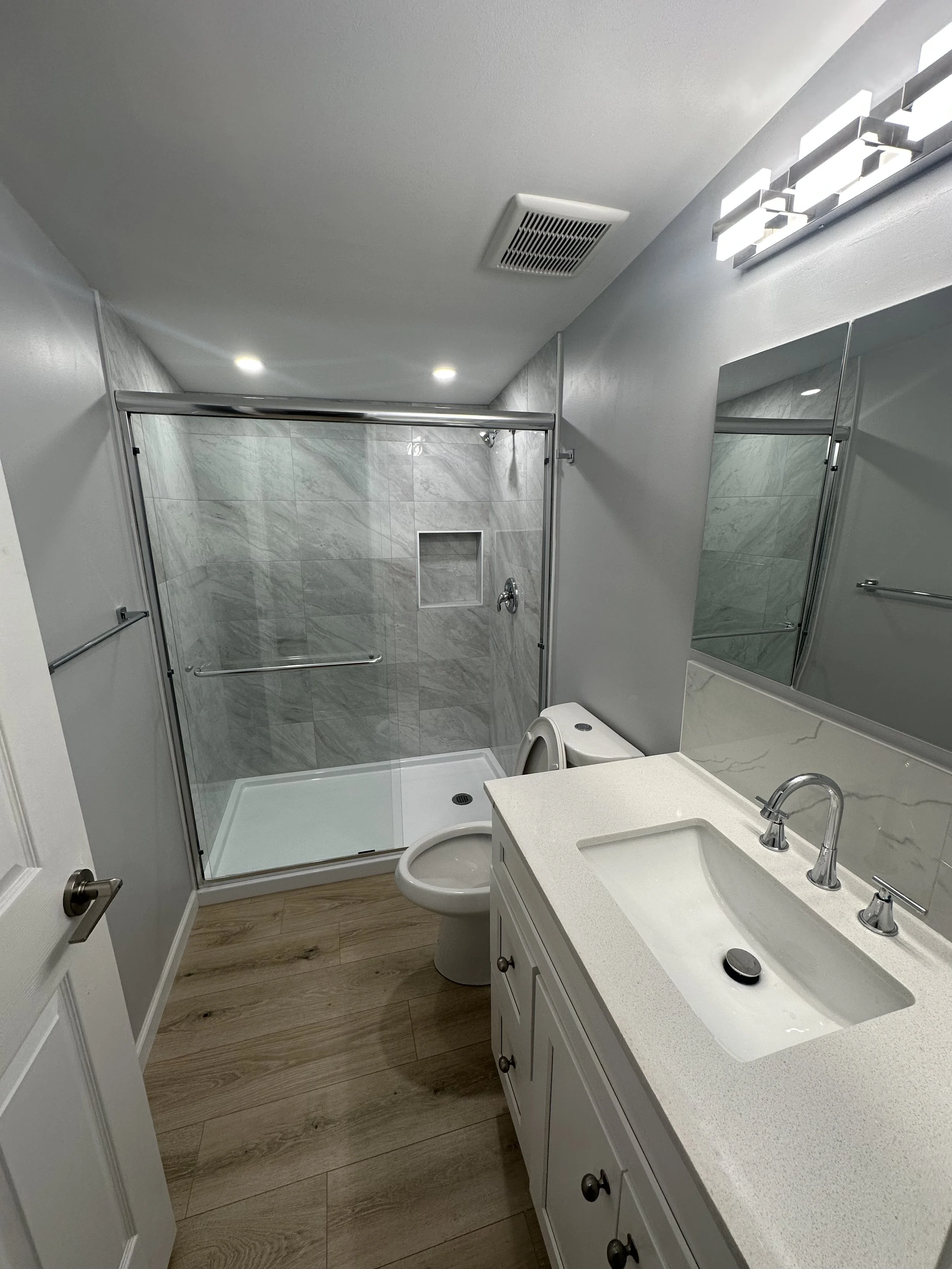 A modern bathroom with a white vanity and sink, a toilet, a glass-enclosed shower with marble tiles, a mirror, and wall-mounted lighting fixtures.
