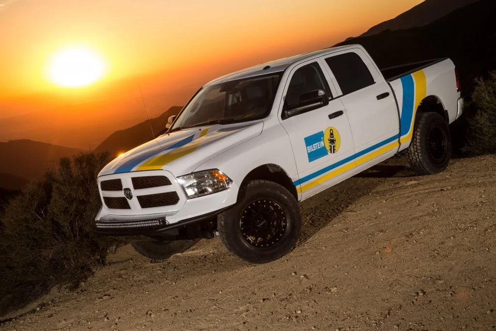 A white pickup truck with Bilstein and other racing decals parked on a dirt trail during sunset, with mountains in the background.