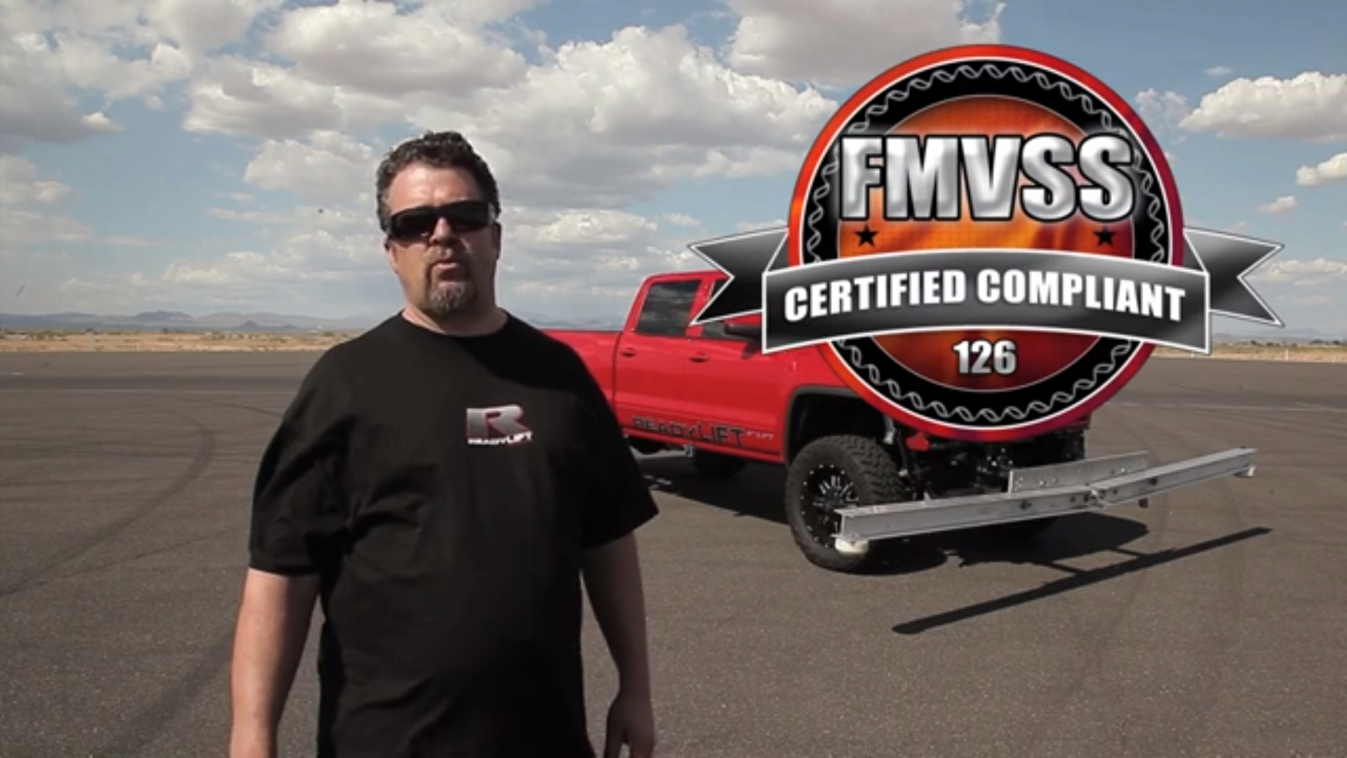 A man wearing sunglasses and a black t-shirt standing on a wide open paved area with a red truck and a large certification badge overlayed on the image. The badge reads 'FMVSS Certified Compliant 126'.