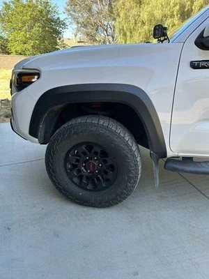 Close-up of the front driver side of a white pickup truck with a black tire and wheel, parked on a concrete driveway with trees in the background.