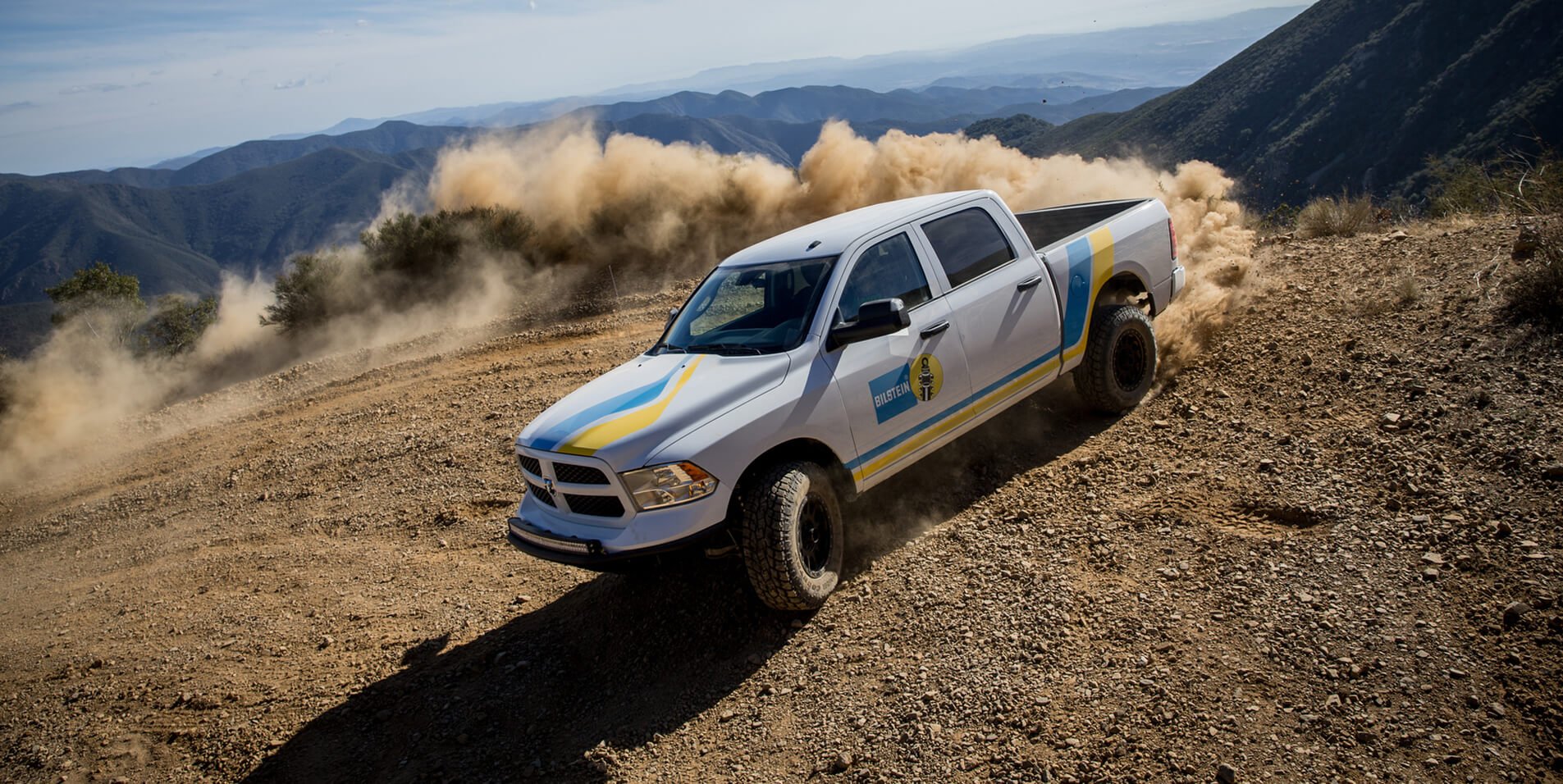 A white off-road pickup truck driving on a dirt trail in a mountainous area, kicking up dust behind it.