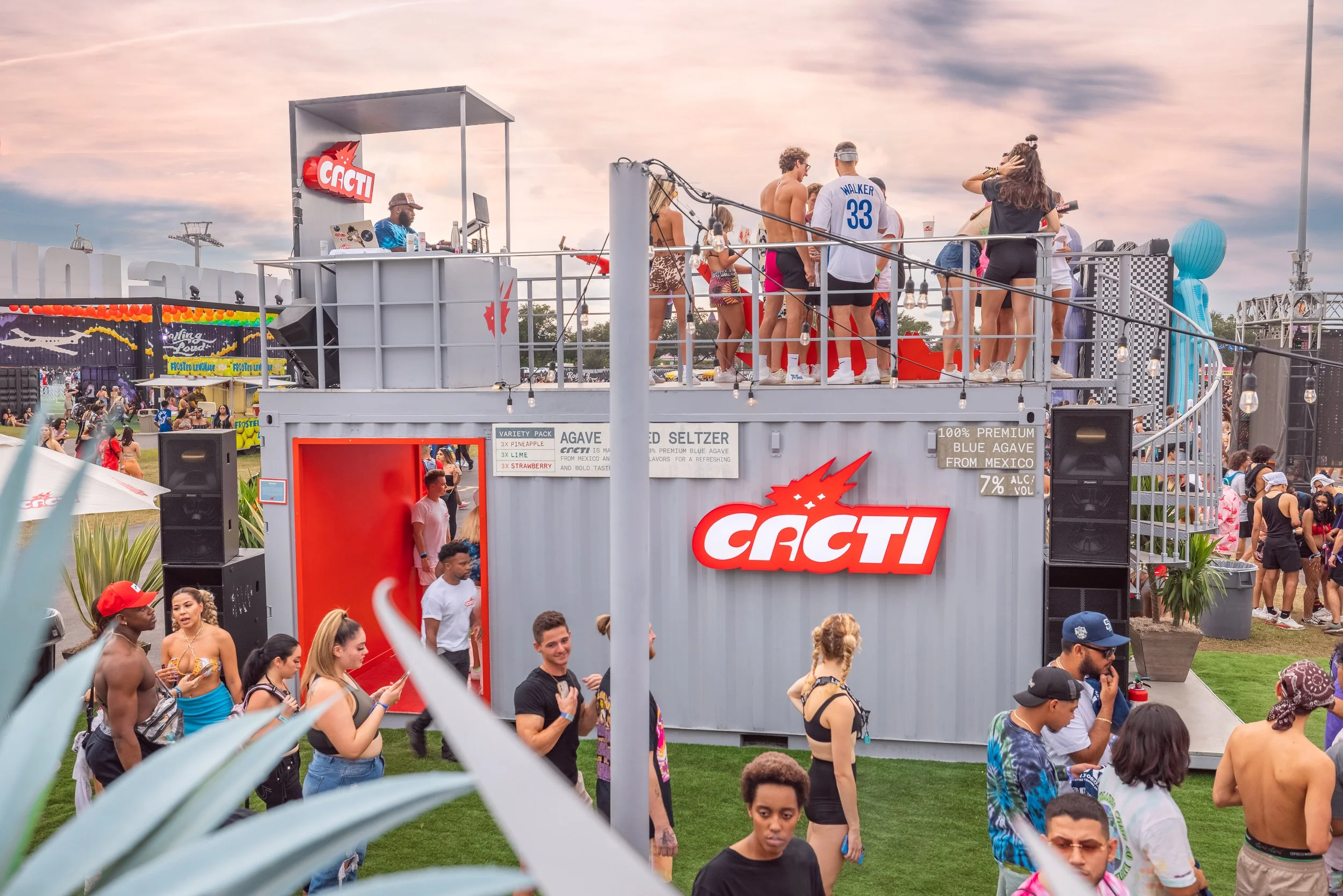 People at an outdoor event, some on a raised platform with DJ equipment and others walking on grass, with tents and rides in the background during sunset.