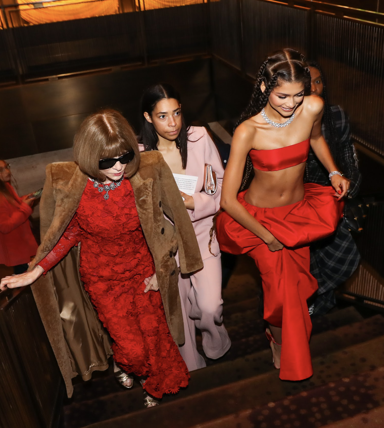 A group of stylish women walking up a staircase at an indoor event, wearing fashionable clothing, including a woman in a bold red dress with an exposed midriff, and another in a fitted red lace dress with a brown coat, surrounded by others in fashion