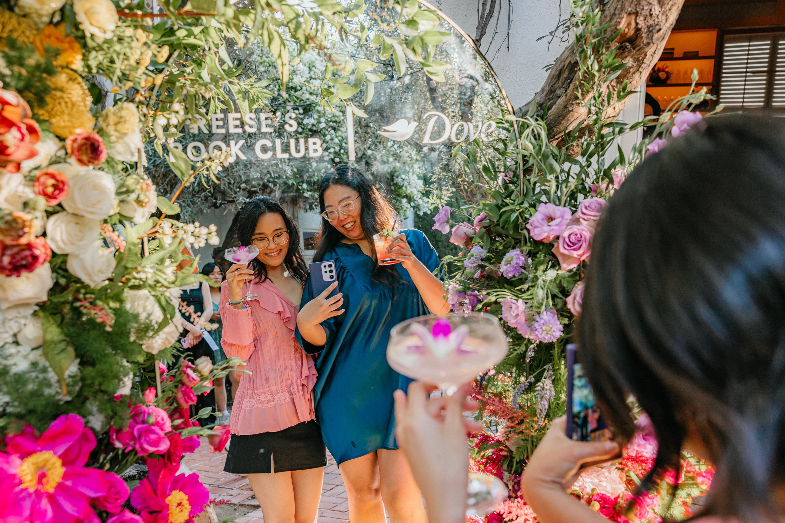 Two women are taking a photo together at an outdoor garden event with vibrant flowers and greenery. One woman holds a drink and a phone, smiling for the photo, while another woman holds a wine glass with a pink flower garnish. The scene is lively and