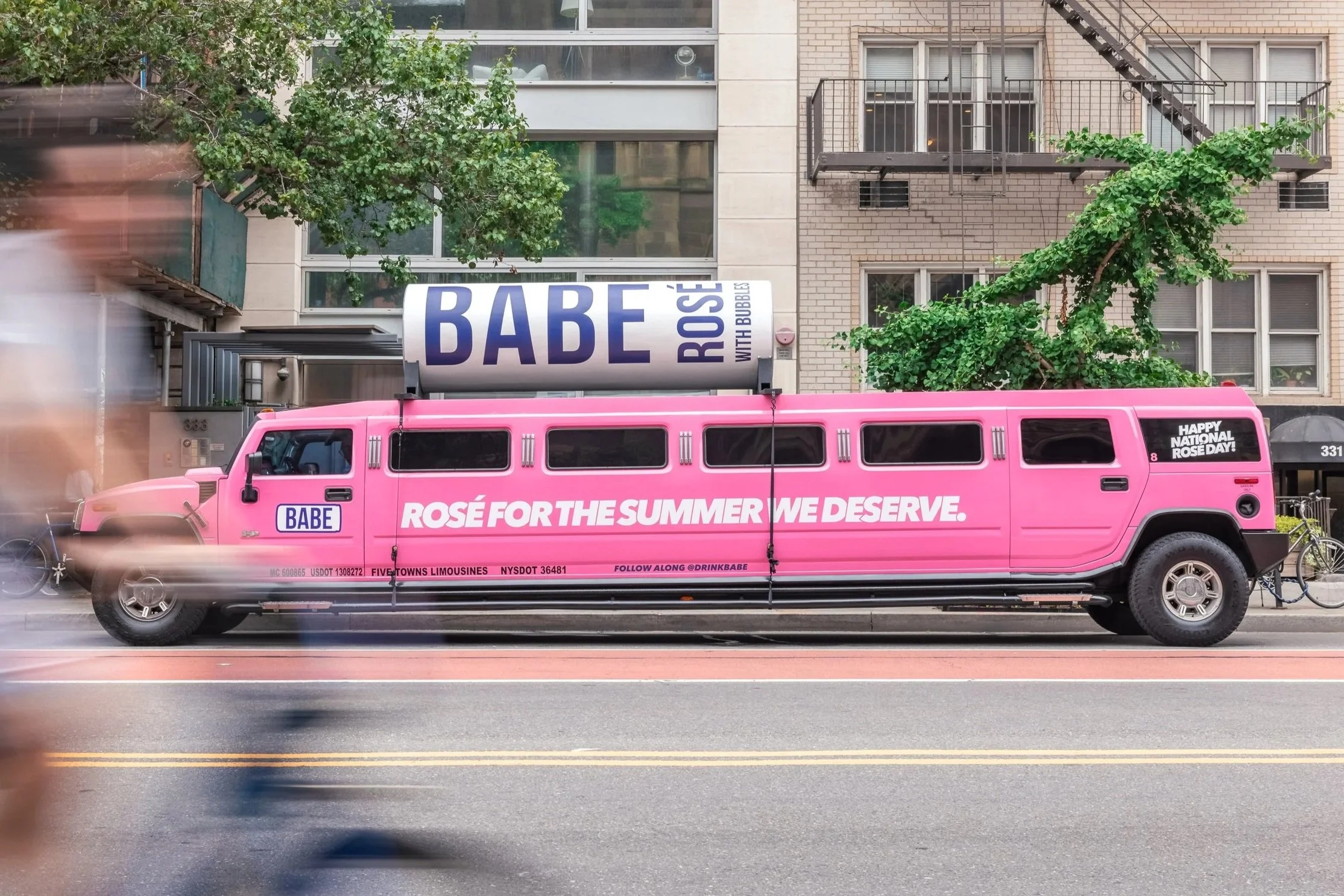 Pink limousine with promotional messages parked on a city street, with blurred pedestrian in the foreground, residential buildings, trees, and a canopy in the background.