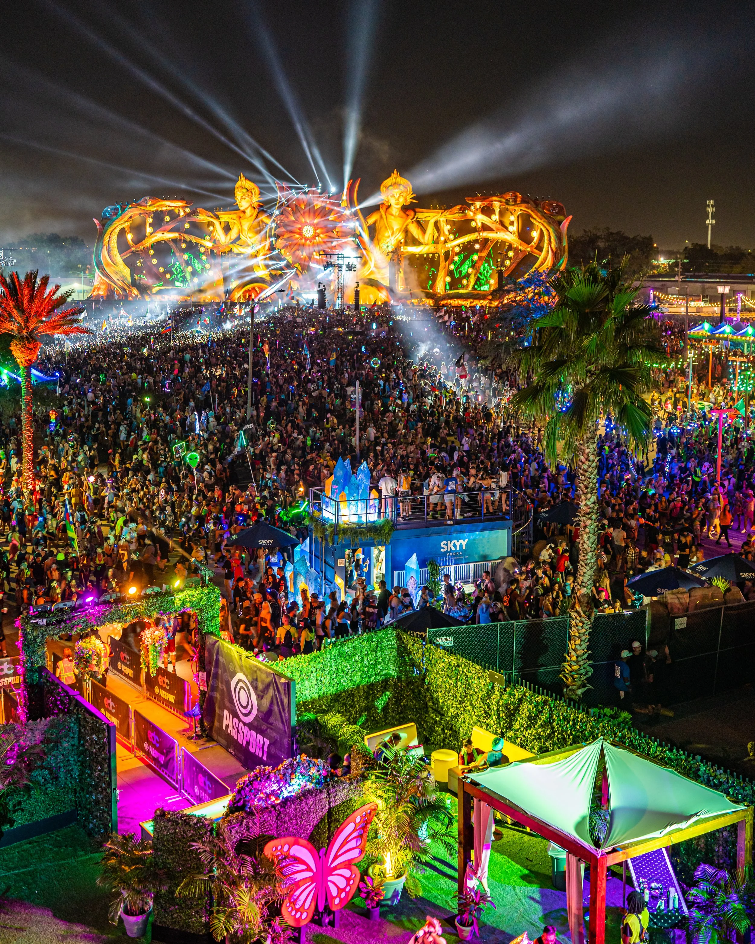 Night scene at a music festival with large, illuminated stage featuring golden figures and beams of light. Crowd gathered in front of stage, colorful decorations, palm trees, and festival booths nearby.