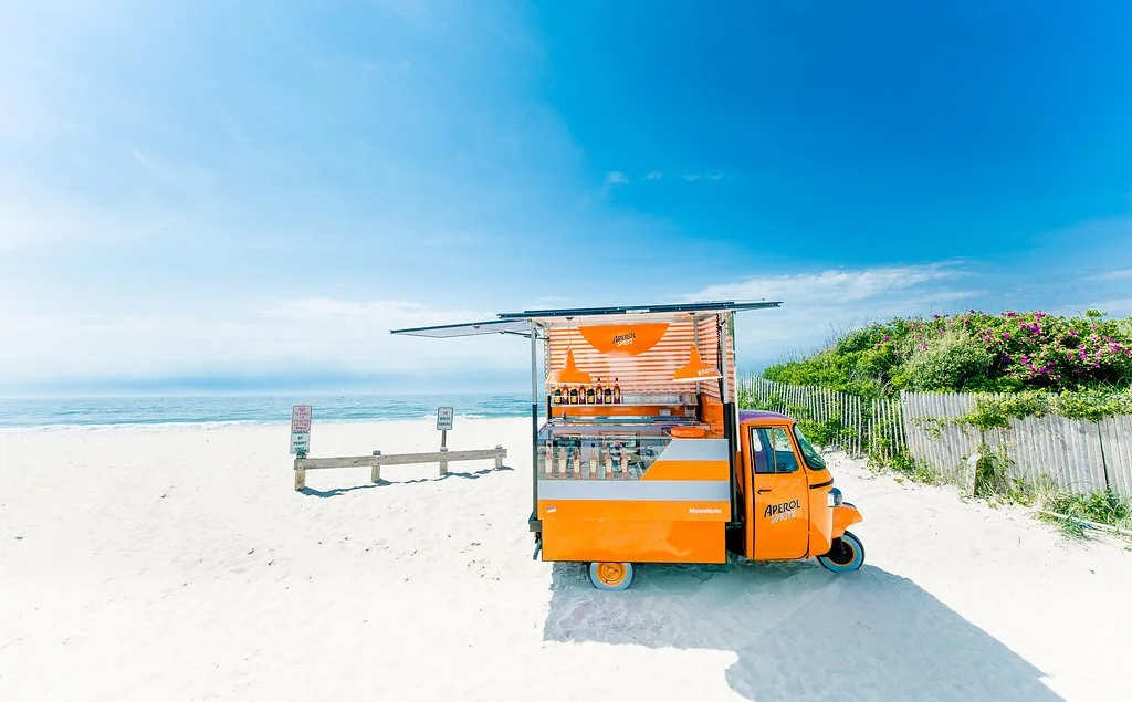 Orange beverage truck with striped awning on a sandy beach, with ocean and blue sky in the background, and greenery on the right.