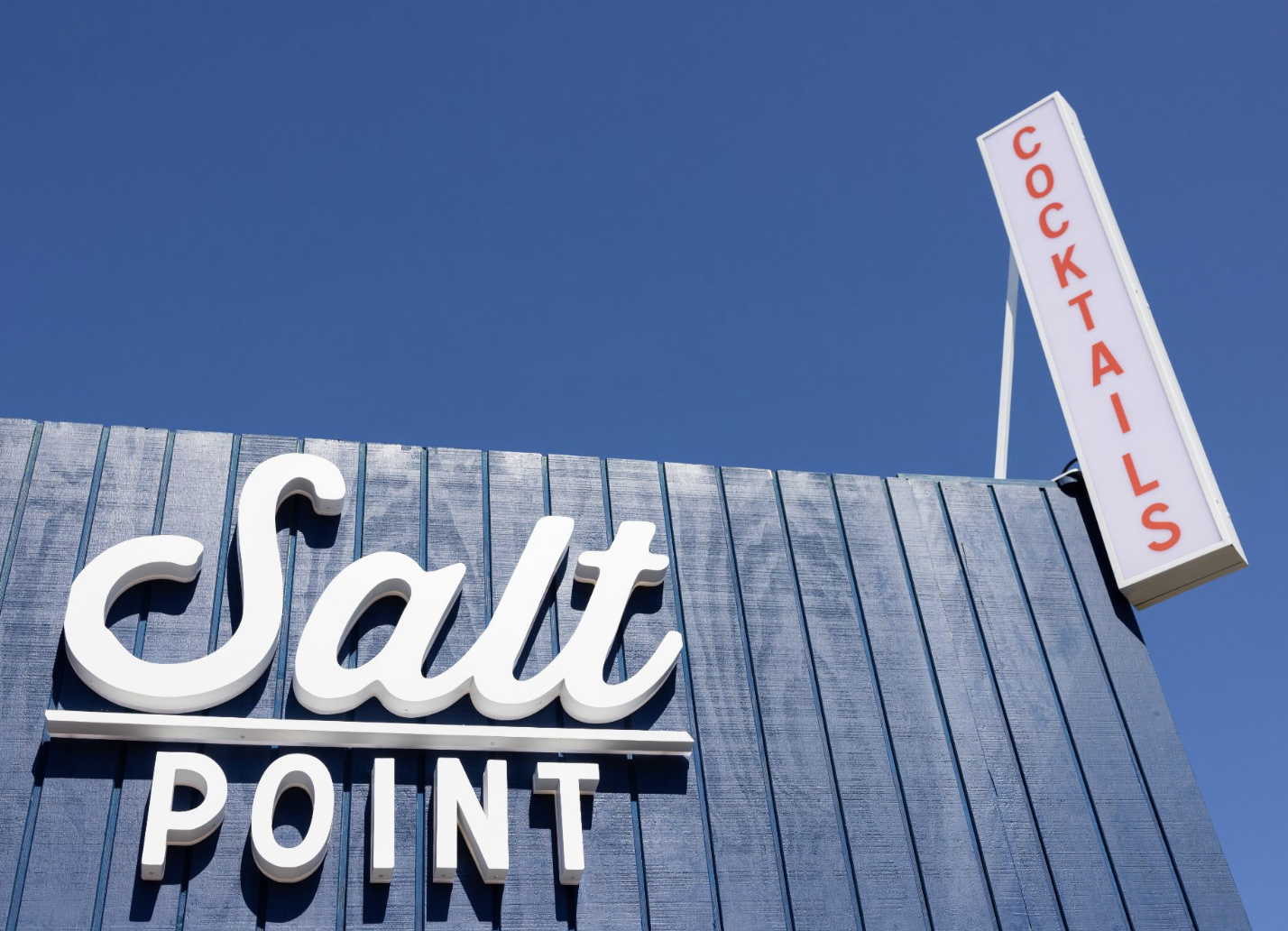 Close-up of Salt Point store sign with white 3D letters on a blue vertical siding background, and a vertical white sign with red letters reading 'COCKTAILS' against a clear blue sky.