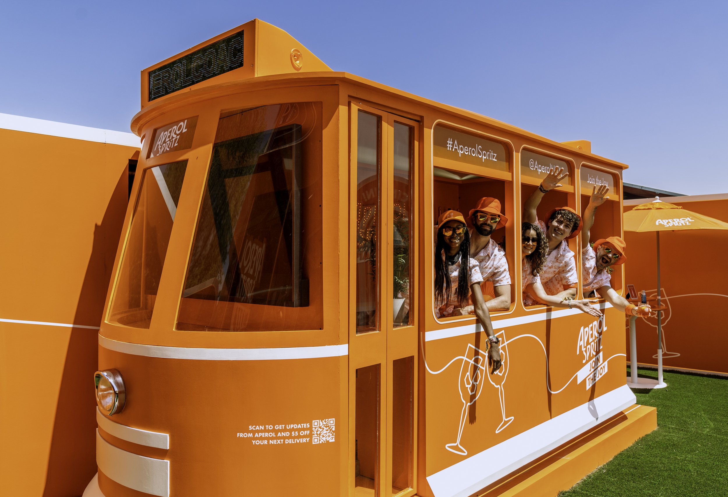 A group of five people enjoying themselves on an orange tram decorated with Aperol Spritz branding, wearing summer outfits and sunglasses, waving and smiling under a blue sky.
