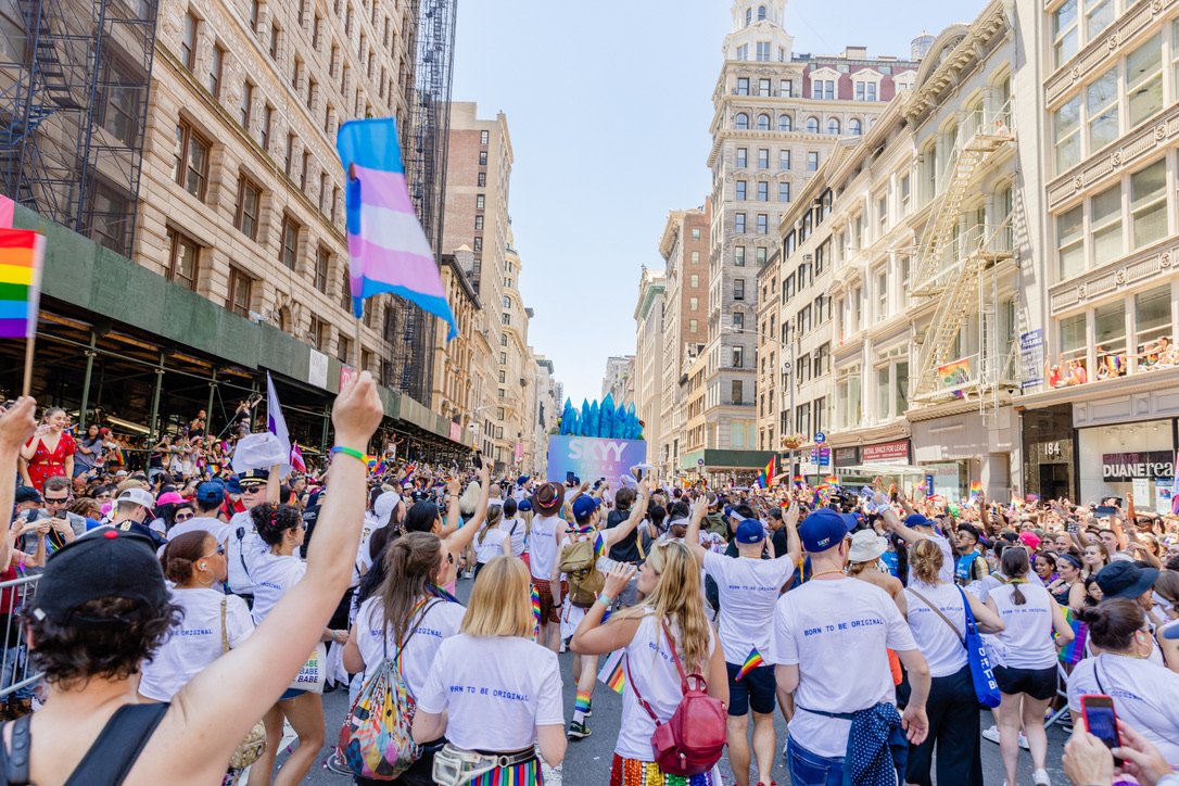 Crowd participating in a Pride parade on a city street with tall buildings, displaying rainbow flags and rainbow-themed clothing.