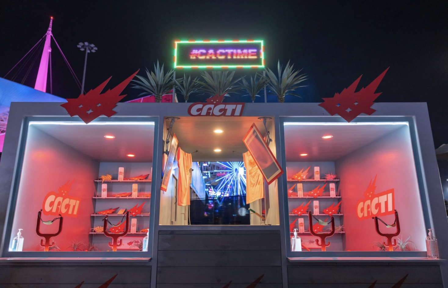 Nighttime at a carnival with a game booth labeled 'Cacti', decorated with red cactus graphics, palm trees, and an LED sign reading '#Cacti', with lights and a ferris wheel in the background.