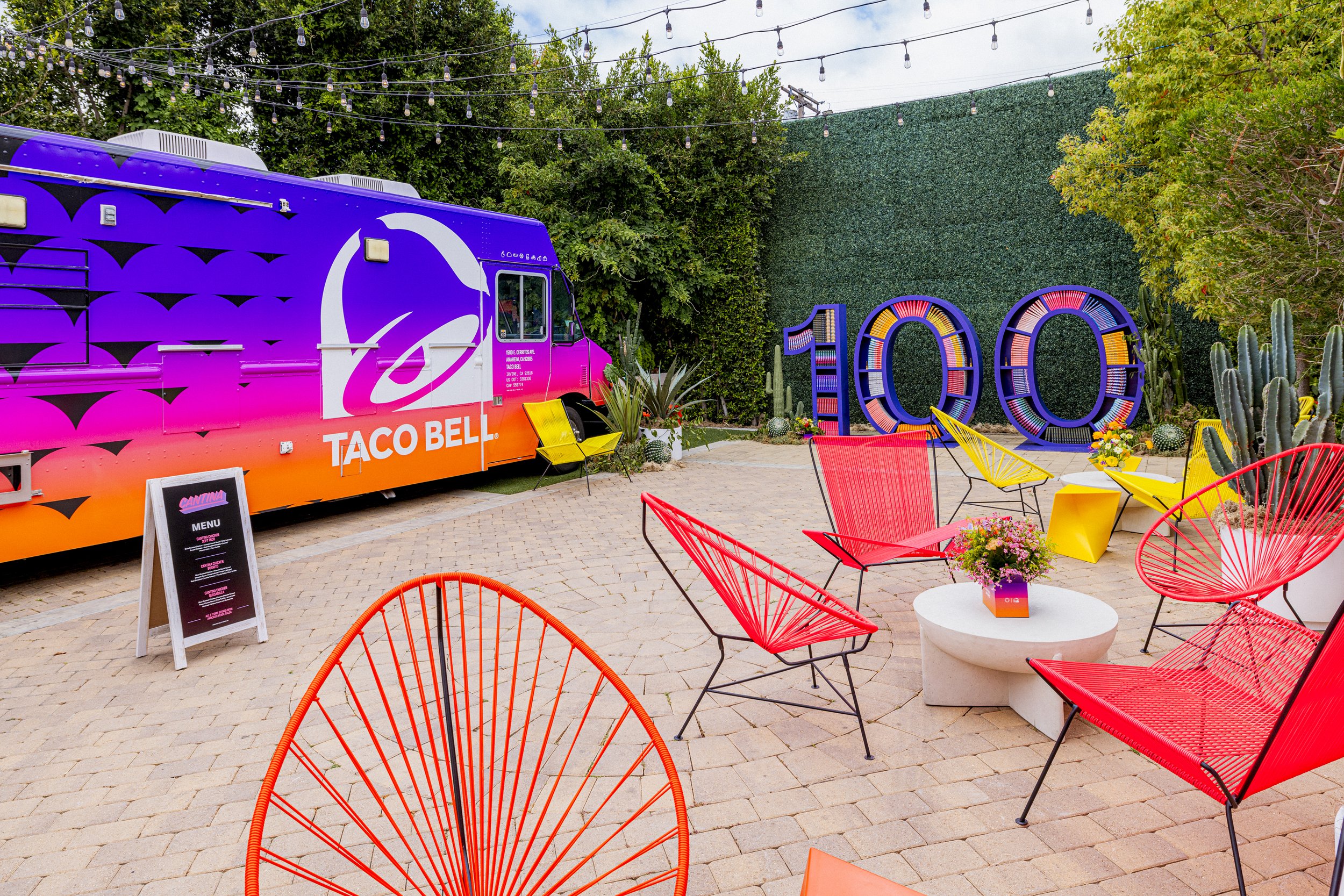 Colorful outdoor patio with yellow and red chairs, a white round table with a flower arrangement, a Tie Belt food truck, and decorative large number 100 against a green hedge backdrop.