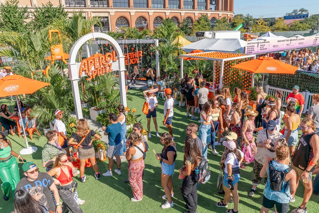 People gathered at an outdoor Apérol Spritz event with orange umbrellas and decorations, surrounded by palm trees and greenery, with a building and tents in the background.