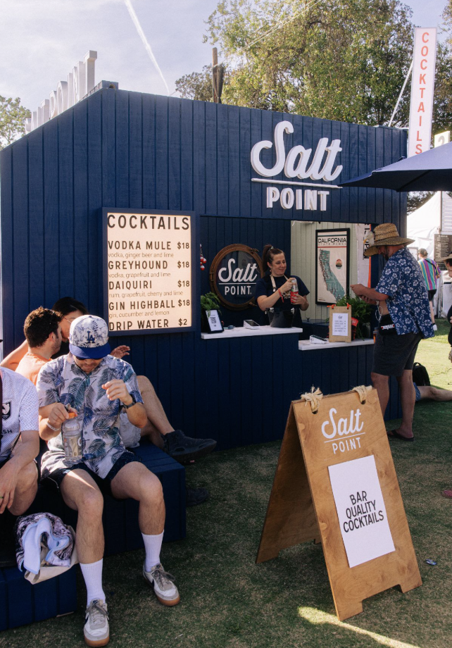An outdoor bar named Salt Point serving cocktails such as vodka mule, greyhound, daiquiri, and gin highball at a festival or event. Customers are ordering and sitting nearby, with a menu sign listing prices and drinks.