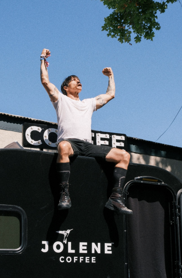 A woman sitting on the roof of a black coffee truck with her arms raised in triumph, wearing a white t-shirt, black shorts, and black boots, with a clear blue sky and a tree branch overhead.