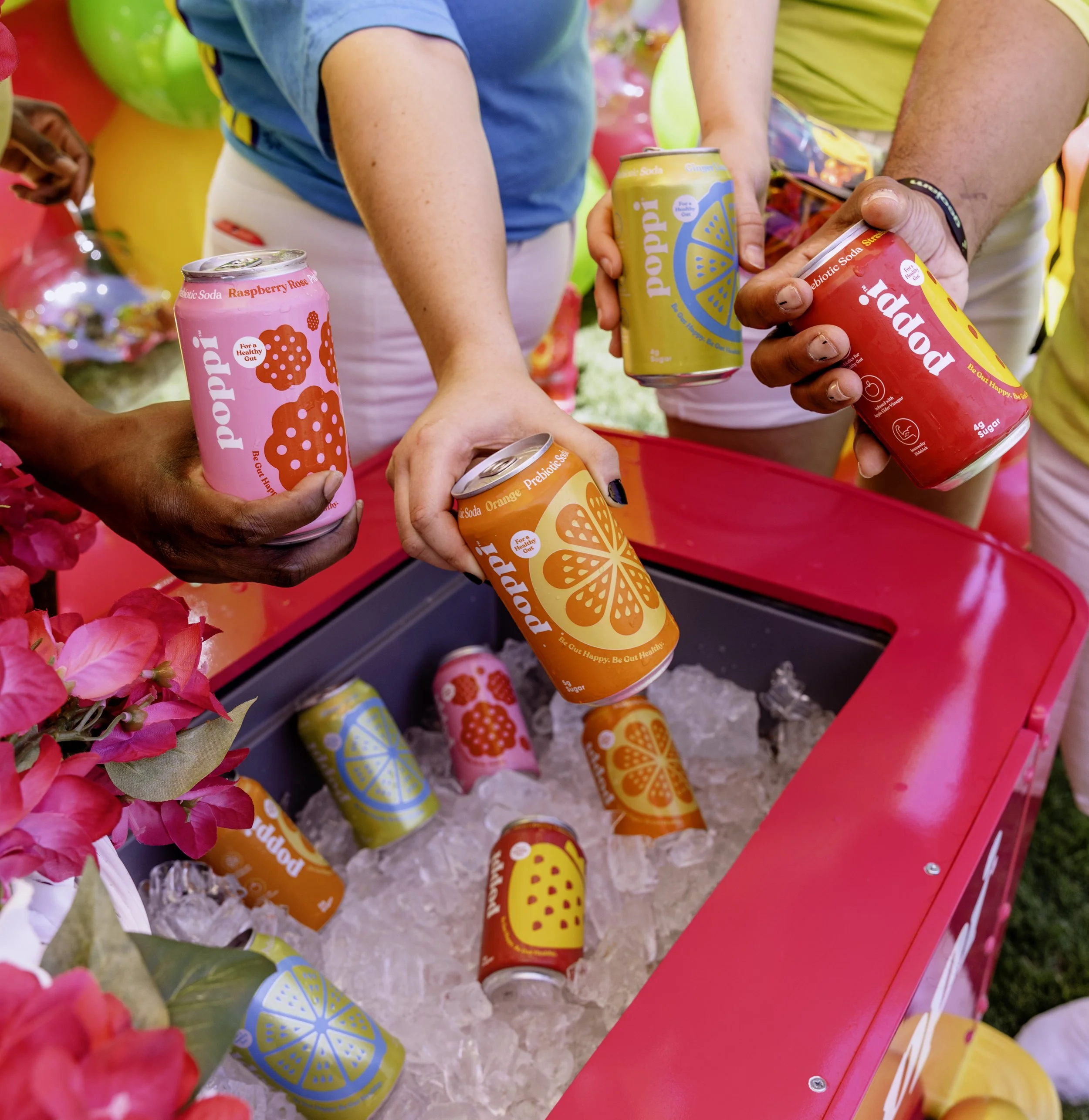 People holding colorful cans of Poppi prebiotic soda over a cooler filled with ice and additional cans, with pink flowers nearby.