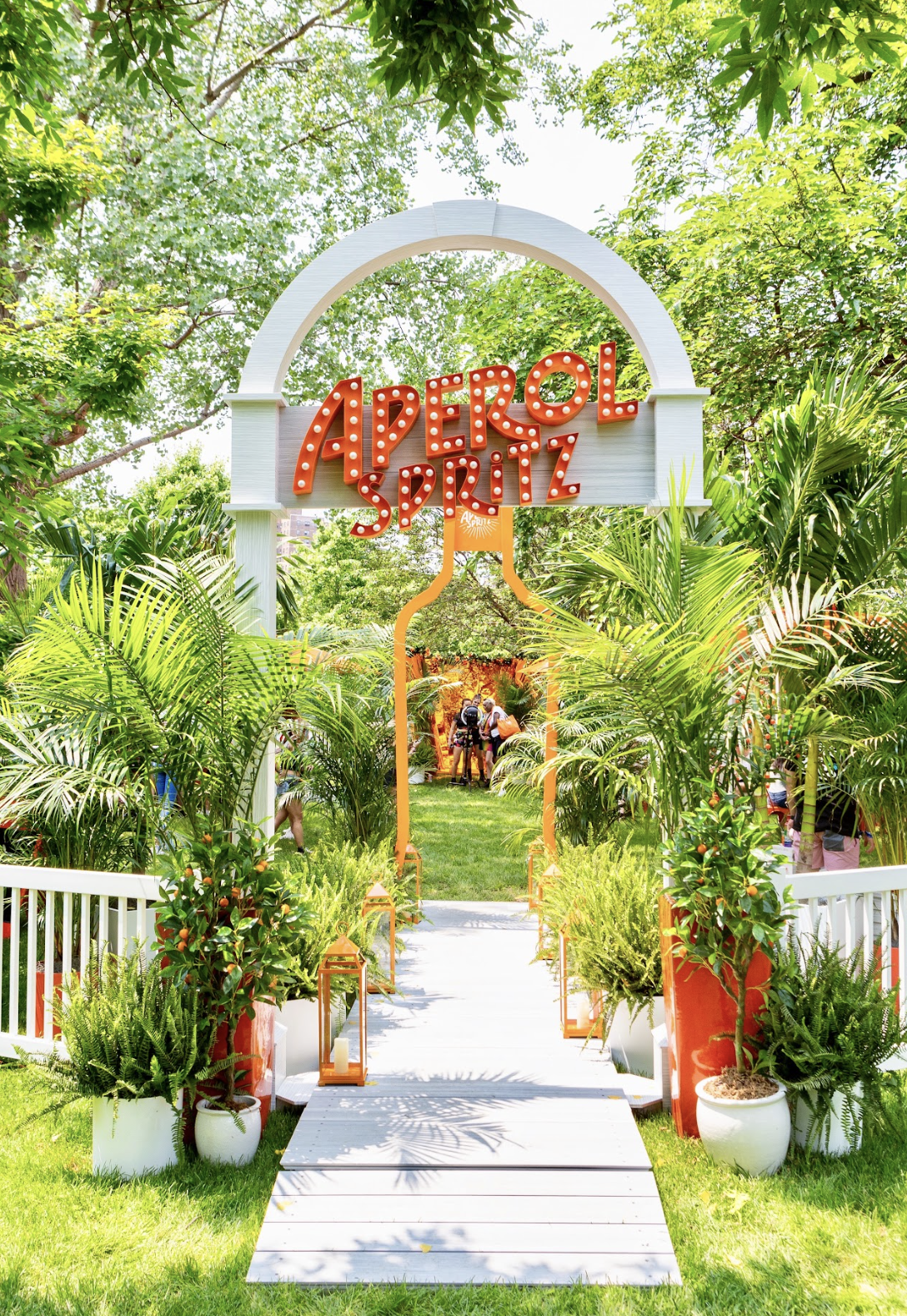 Entrance to an outdoor event called 'Aperol Spritz,' marked by a white archway with a red sign and decorative orange elements, surrounded by lush green plants and trees.