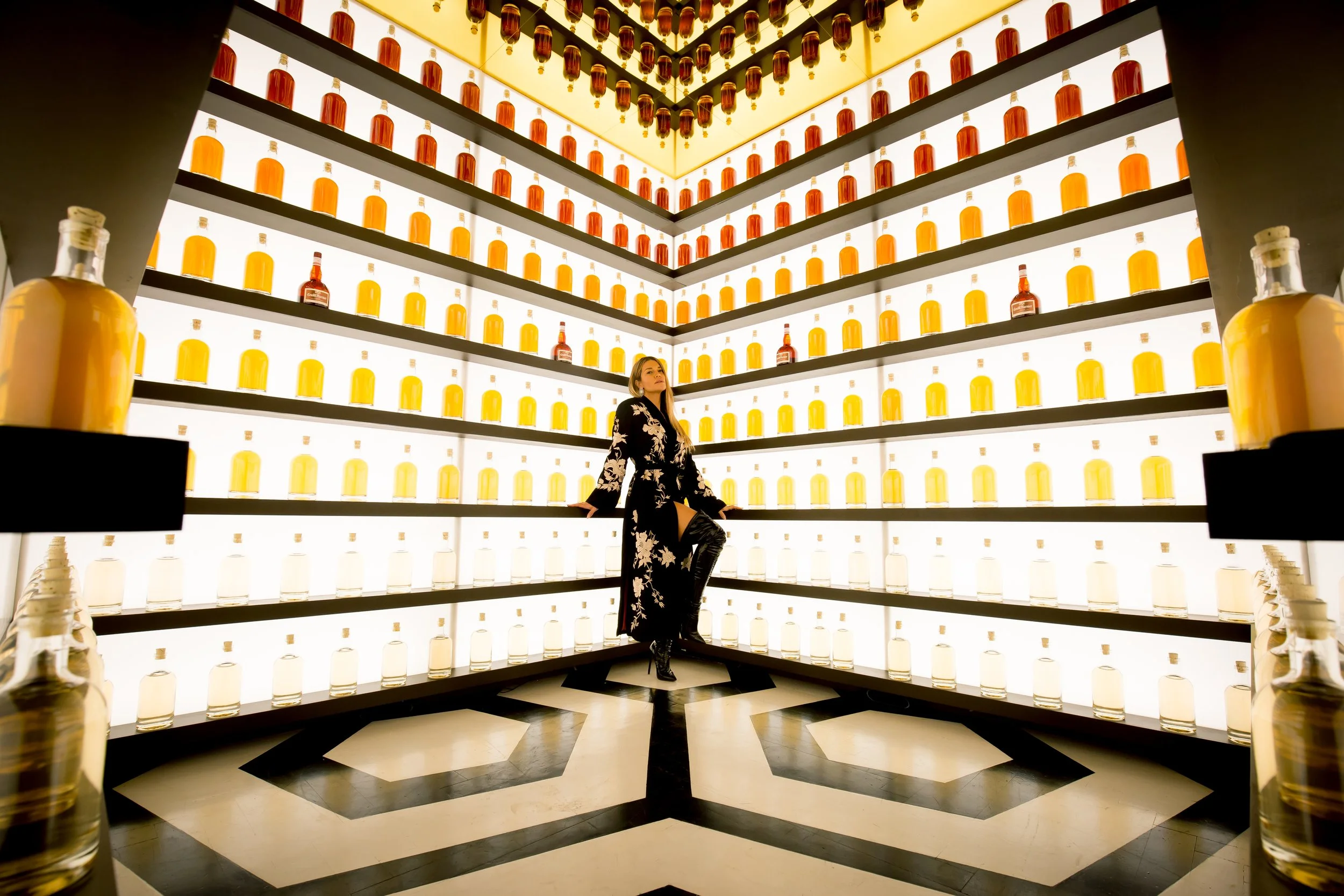 A woman in a black dress with floral pattern and thigh-high boots poses in front of illuminated shelves filled with bottles of yellow and amber liquors, in a luxurious bar or display area.