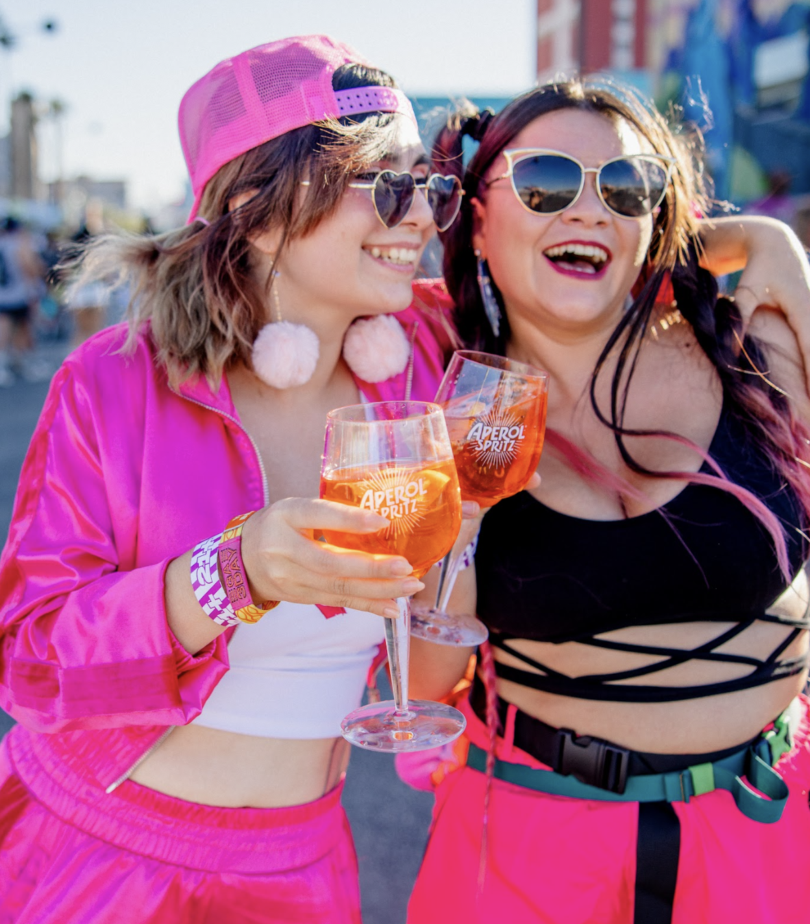 Two women celebrating outdoors, wearing colorful clothing and sunglasses, holding glasses of Aperol Spritz.