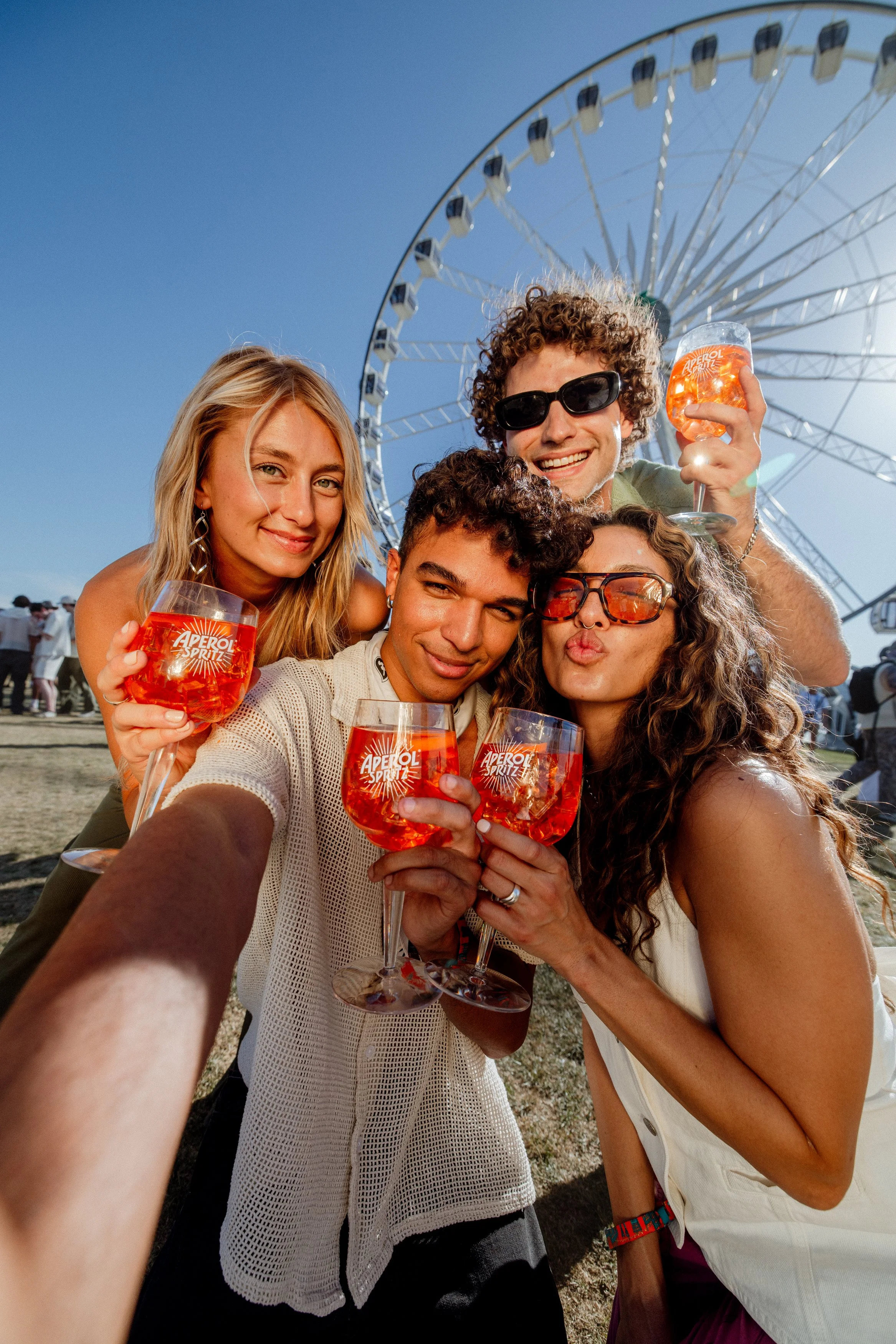 Group of five friends taking a selfie at an outdoor carnival, each holding a glass of Aperol Spritz with a large Ferris wheel in the background, under a clear blue sky.