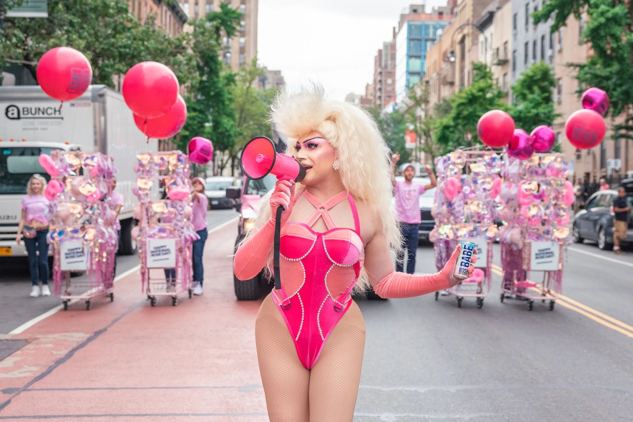 Person with platinum blonde curly hair dressed in a pink outfit speaking into a megaphone on a city street during a parade with balloons and individuals in pink outfits in the background.