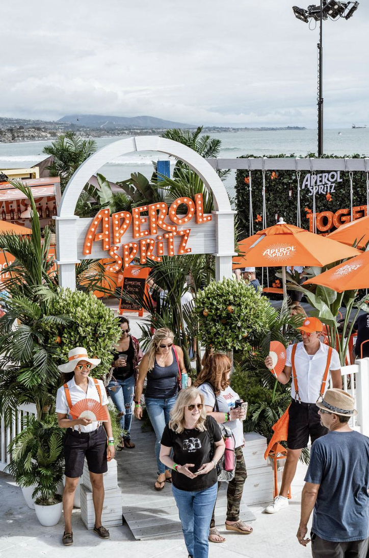 People gather at an outdoor event with large orange umbrellas and a sign reading 'Aperol Spritz'. The setting appears to be near a seaside with a view of the ocean and mountains in the background. The decor features tropical plants, and some people a