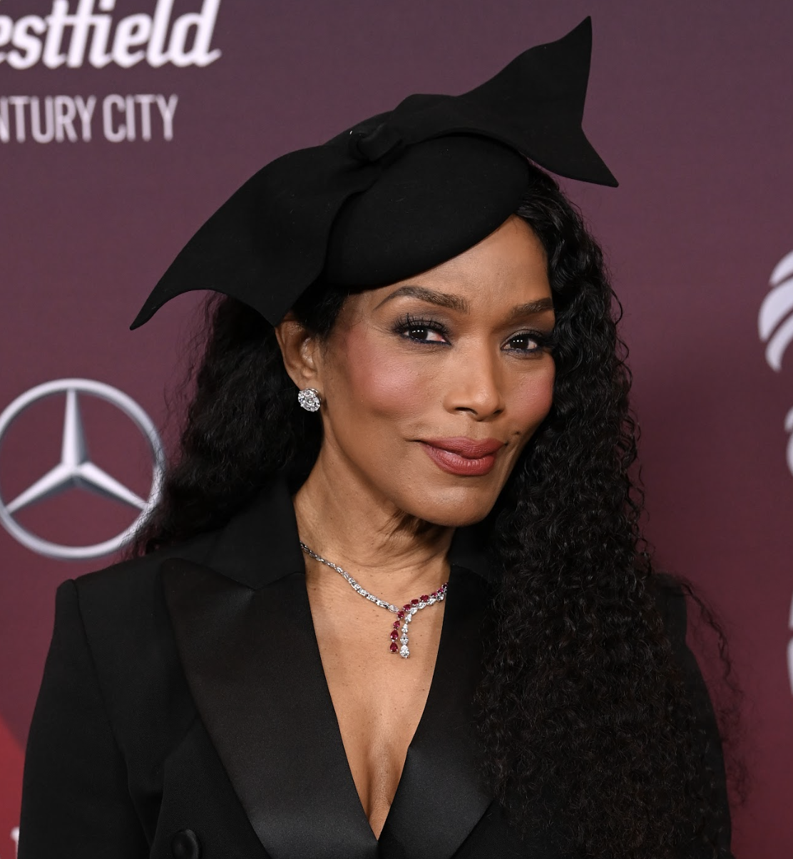 A woman with curly black hair, wearing a black dress or blazer with a V neckline, and a large black hat with a bow, standing in front of a burgundy background with logos and text.