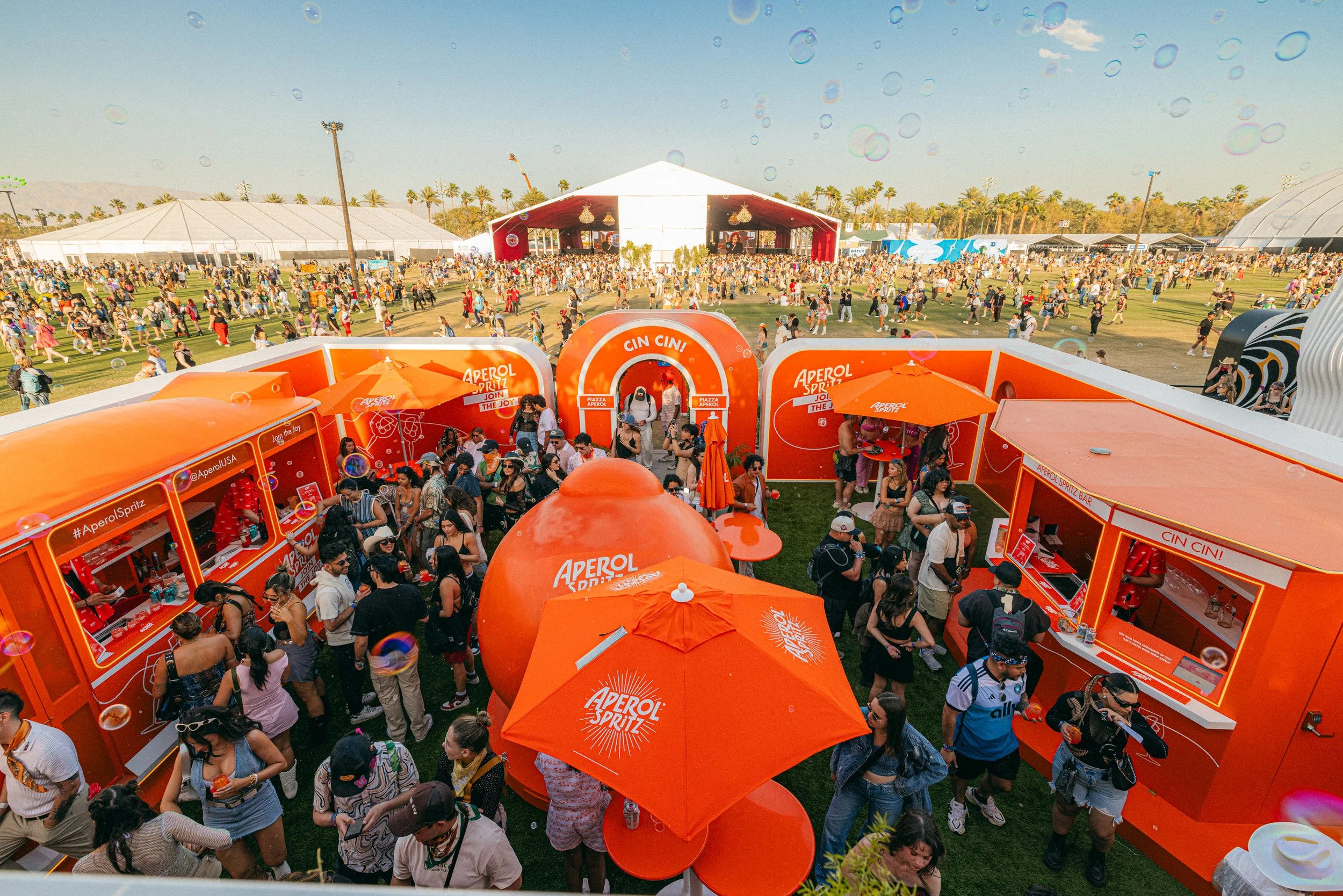 A crowded outdoor festival with a large orange Aperol Spritz bar and multiple smaller booths, surrounded by numerous attendees. The scene includes tents, a stage in the background, and palm trees in the distance, under a clear sky.
