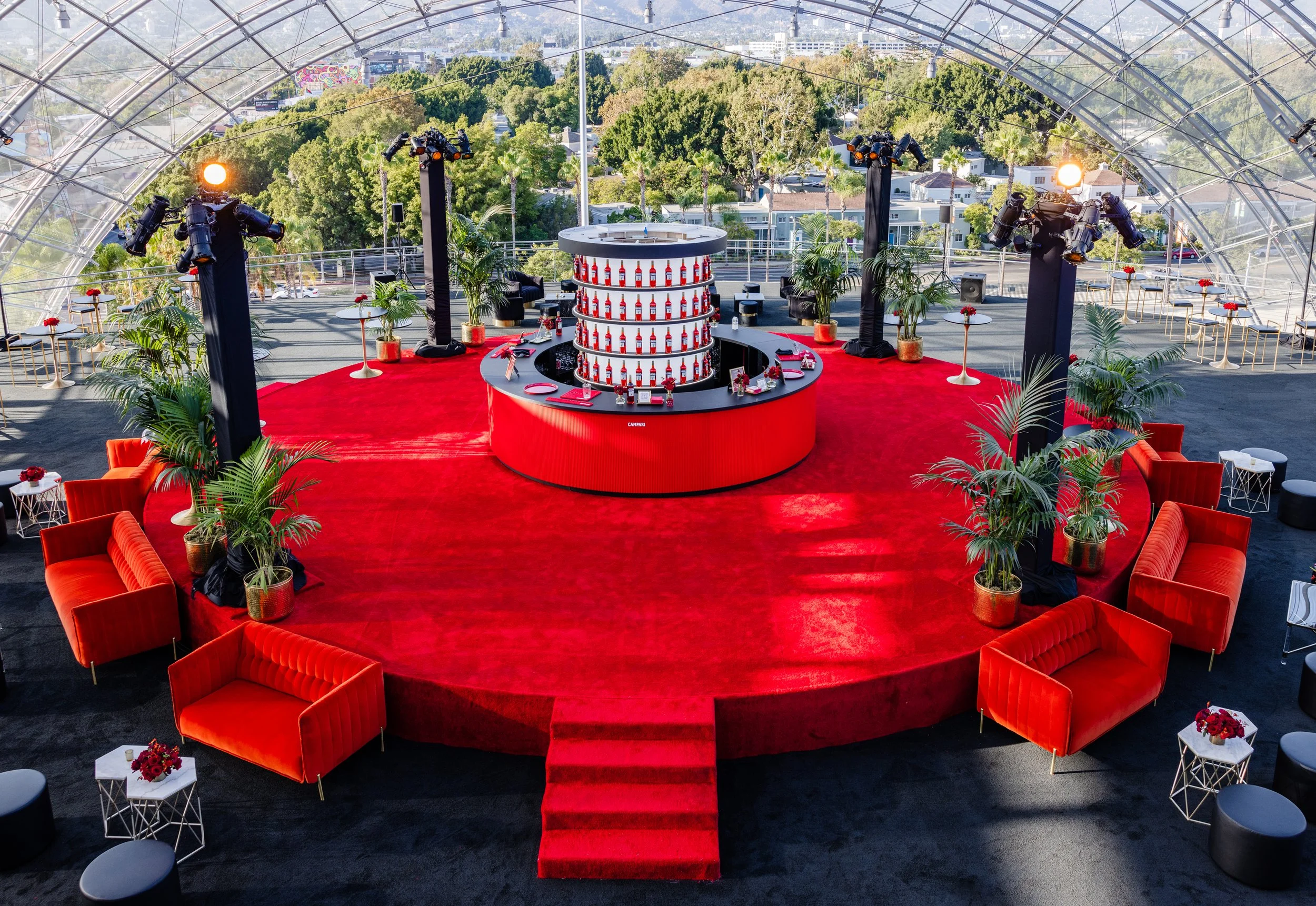 Empty event stage with red carpet and seating area, surrounded by potted plants, tables, and chairs under a transparent dome ceiling with cityscape in the background.