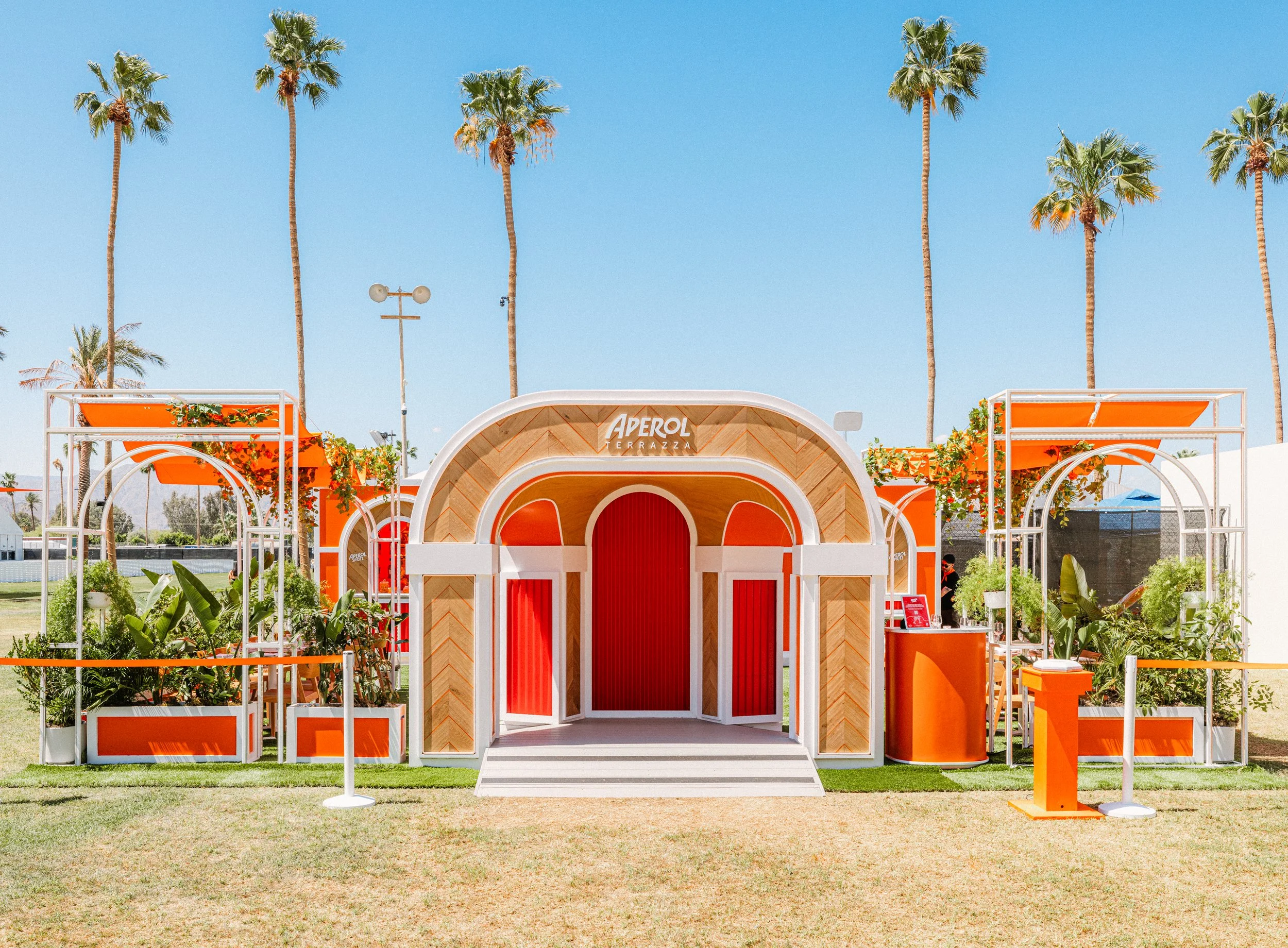 Outdoor pavilion named 'Aperol Terrazza' with bright orange and white color scheme, tropical plants, and tall palm trees under a clear blue sky.
