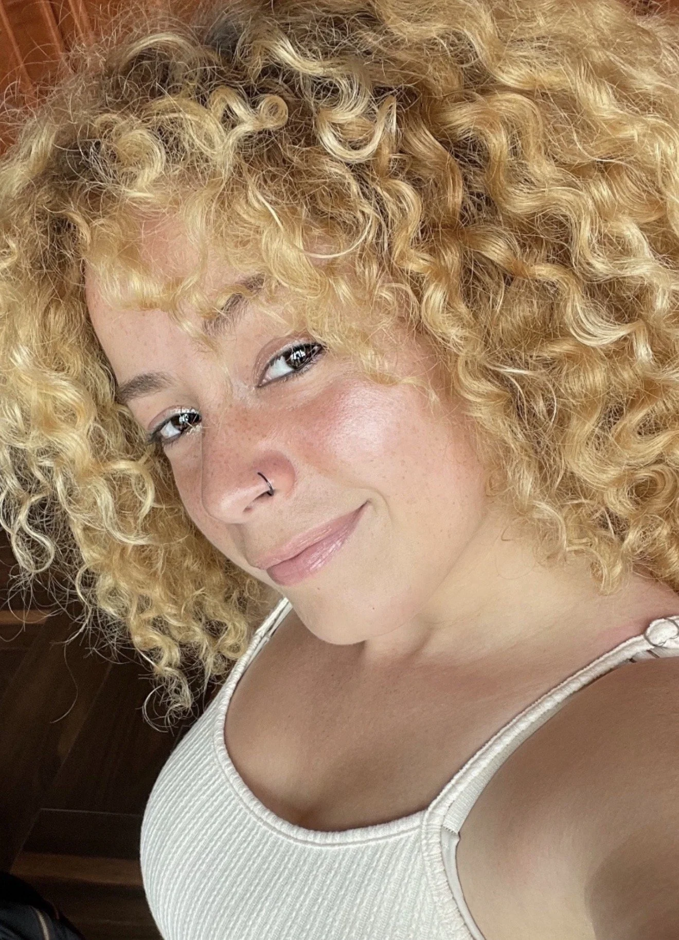 A woman with curly blonde hair, light skin, and freckles taking a close-up selfie, wearing a white tank top.