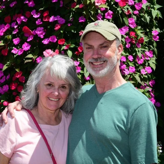A smiling woman and man standing outdoors in front of a bush with pink and purple flowers, with the woman’s arm around the man's shoulders.